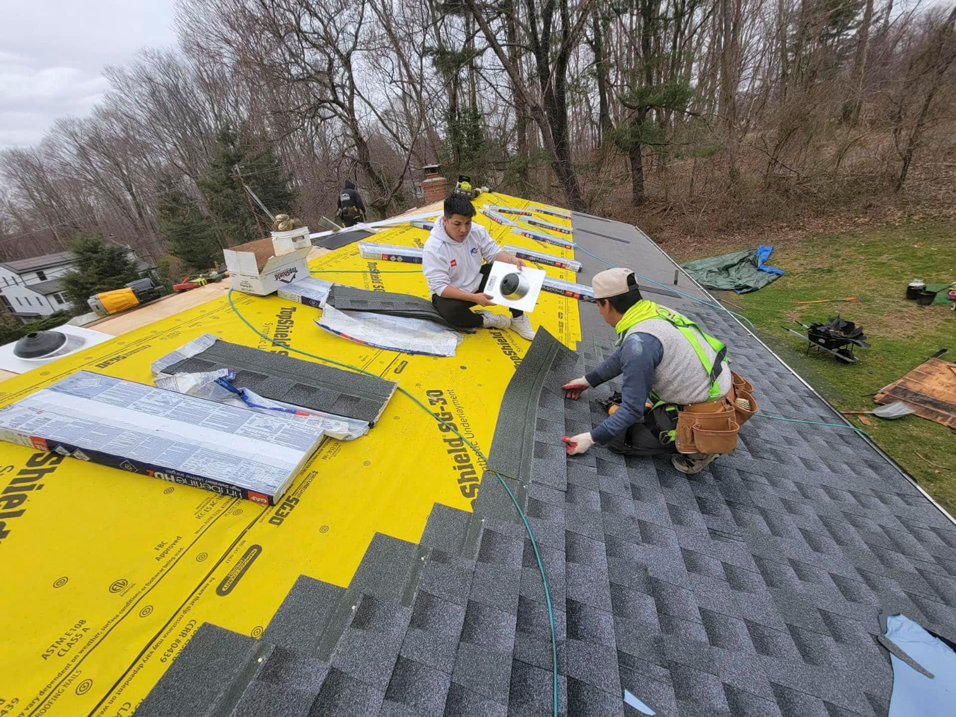 Workers installing shingles on a roof, with gray and bright yellow sections and roofing materials spread out.