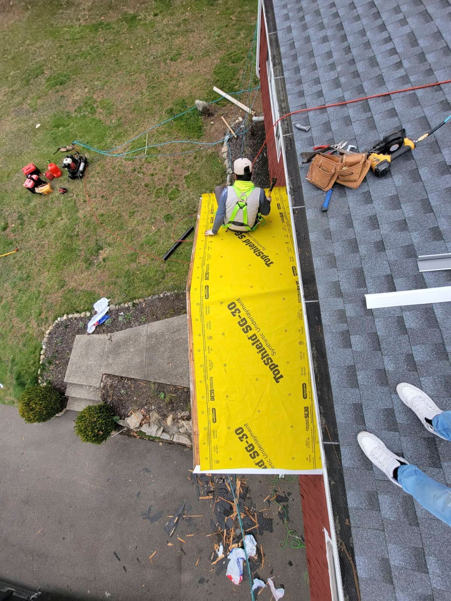 Person on a roof installing bright yellow safety tarp beside a ladder and tools, viewed from above