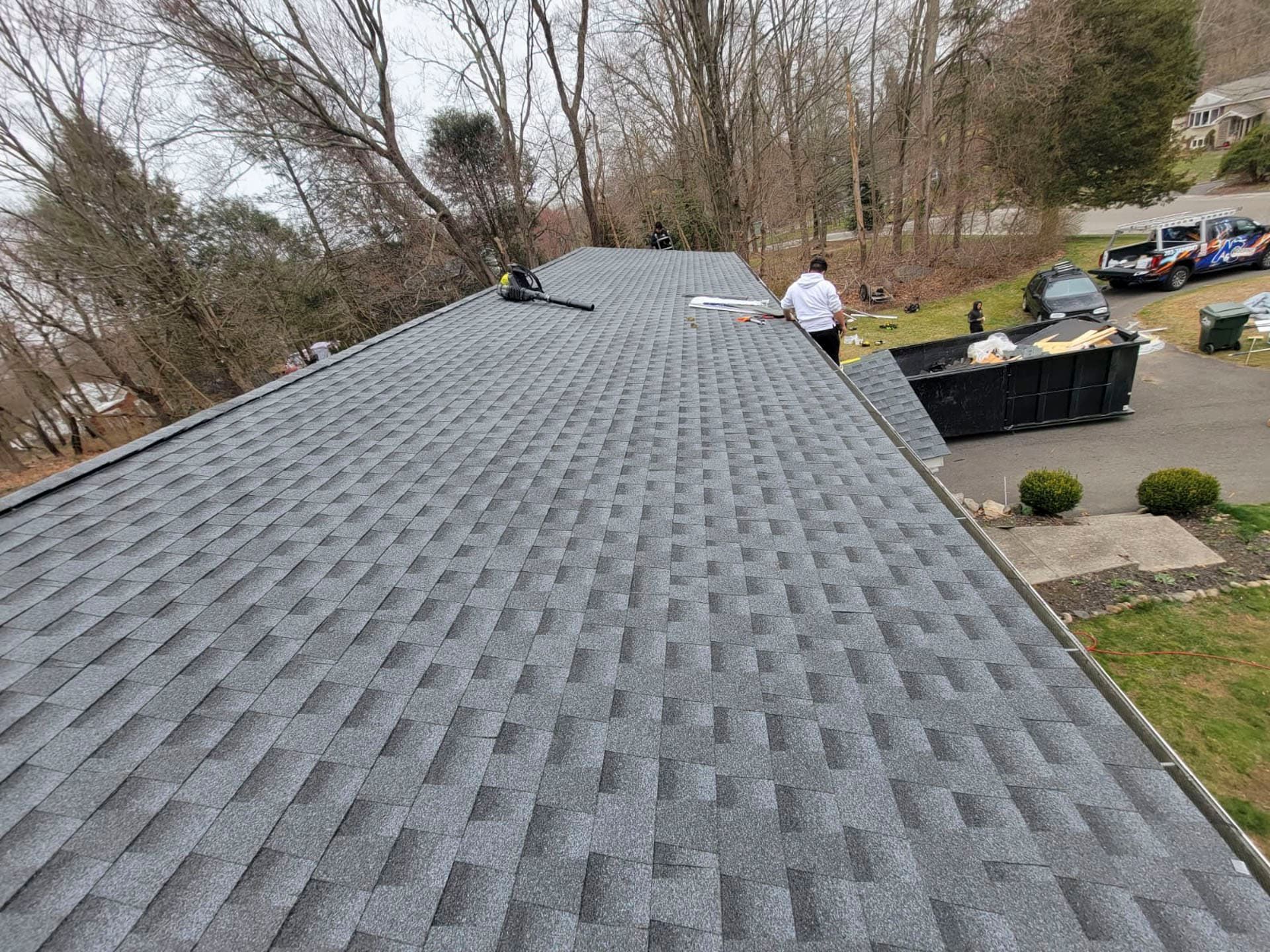 Roof under repair with gray shingles, workers, and black material bins beside a wooded yard