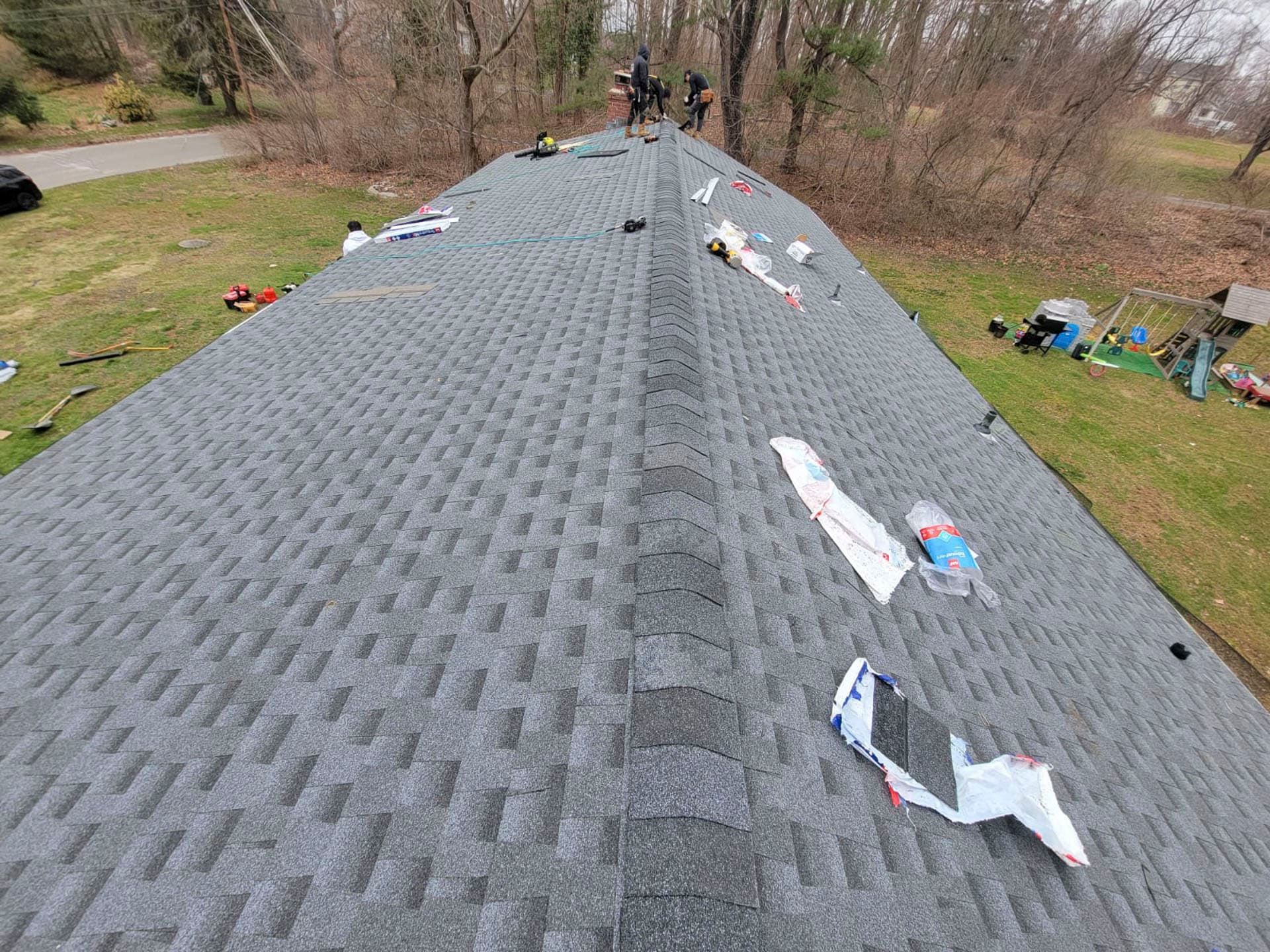 Roof under construction with gray shingles and scattered tools, viewed from the ridge toward the trees below
