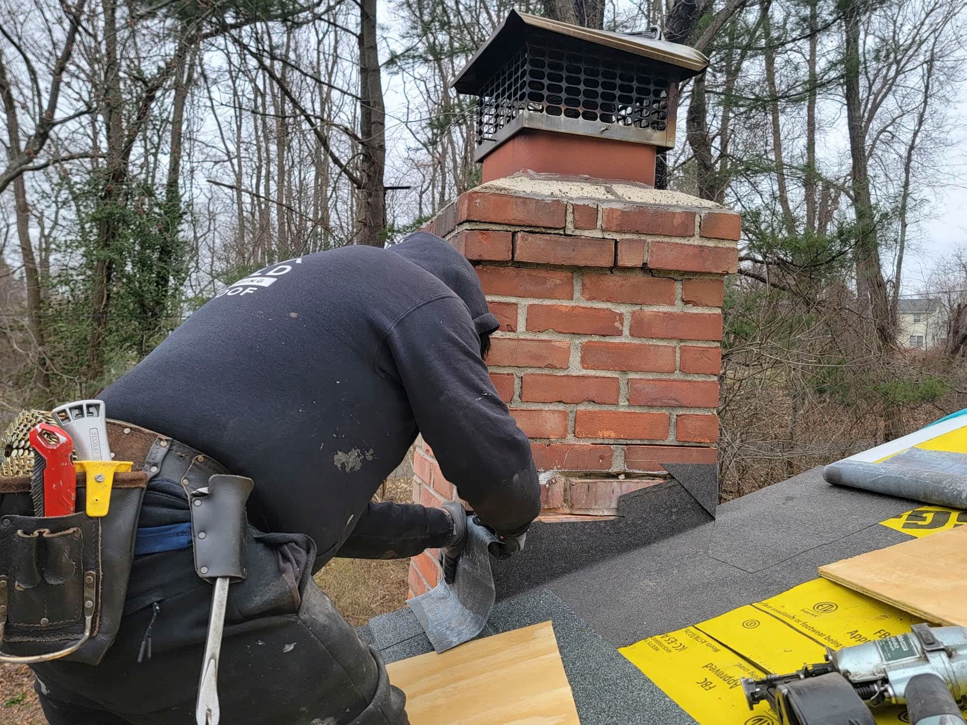 Worker repairing a brick chimney on a shingled roof, with tools and roofing materials nearby.