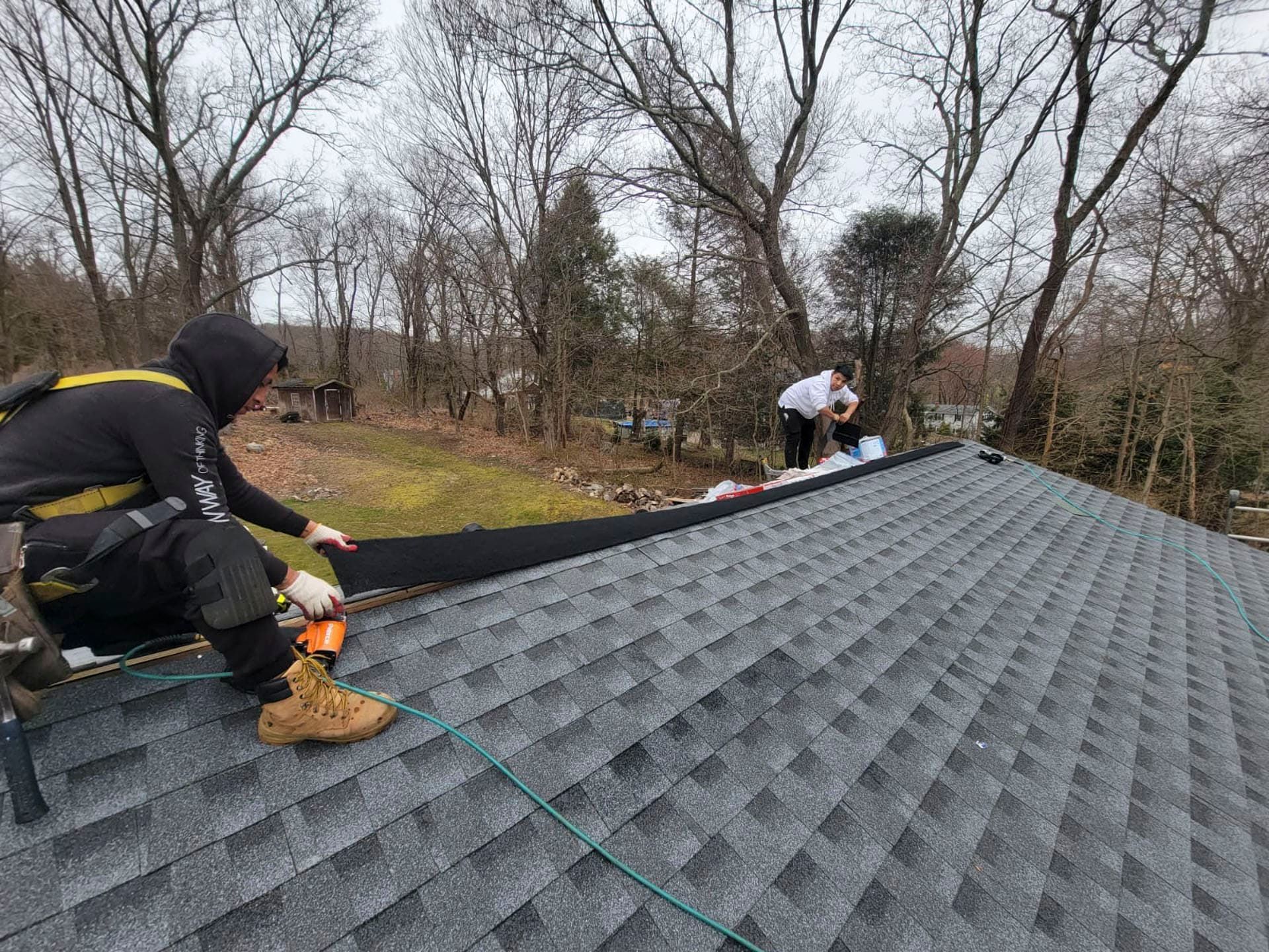 Workers installing gray shingles on a roof in a wooded yard