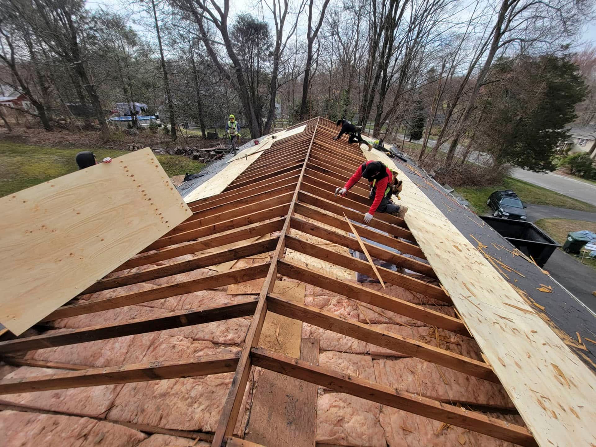 Roofers repairing a house roof, exposed plywood and framing visible on both sides.