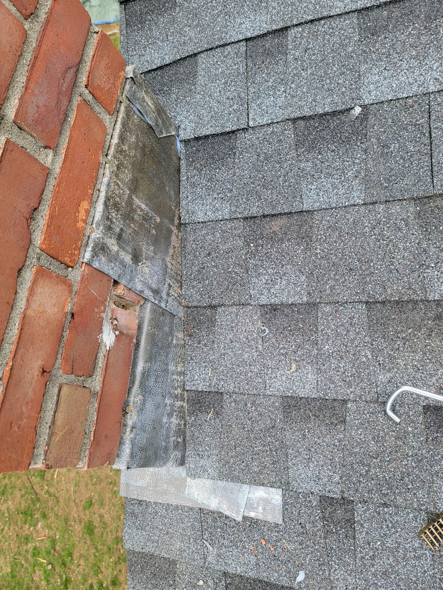 Roof shingles meeting a red brick wall at a corner, with a small patch of grass below.