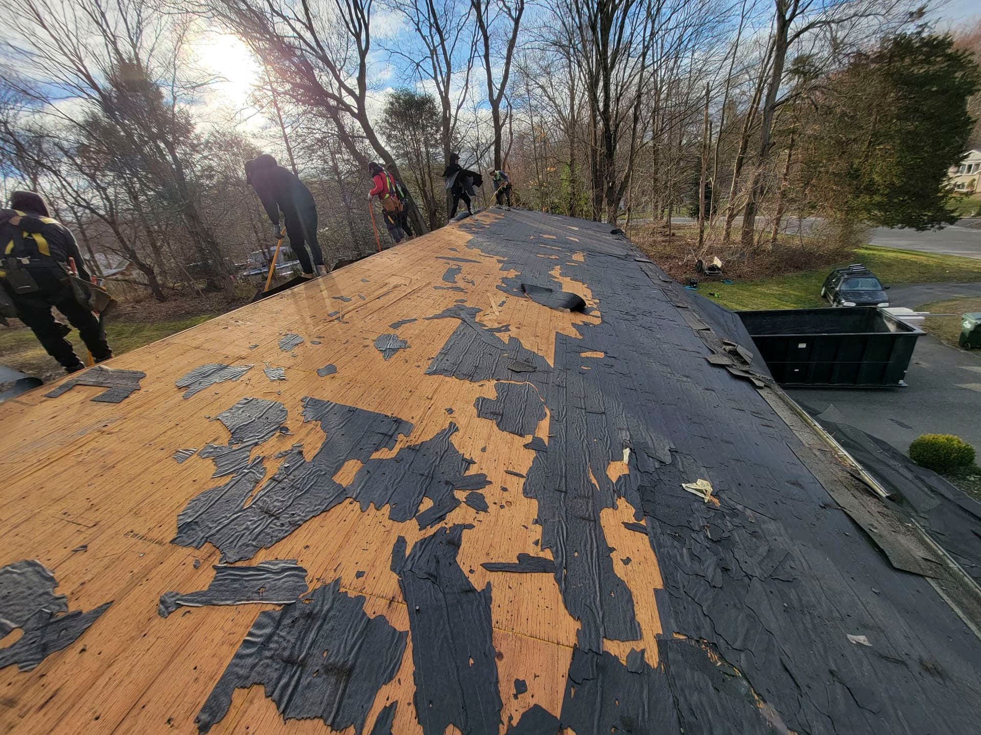 Peeling black roof with orange underlayment, workers in background, trees and bright sun overhead