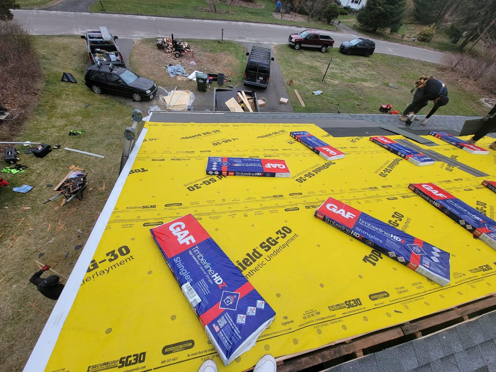 Roofers installing bright yellow roofing underlayment on a house roof, with supplies and tools below.