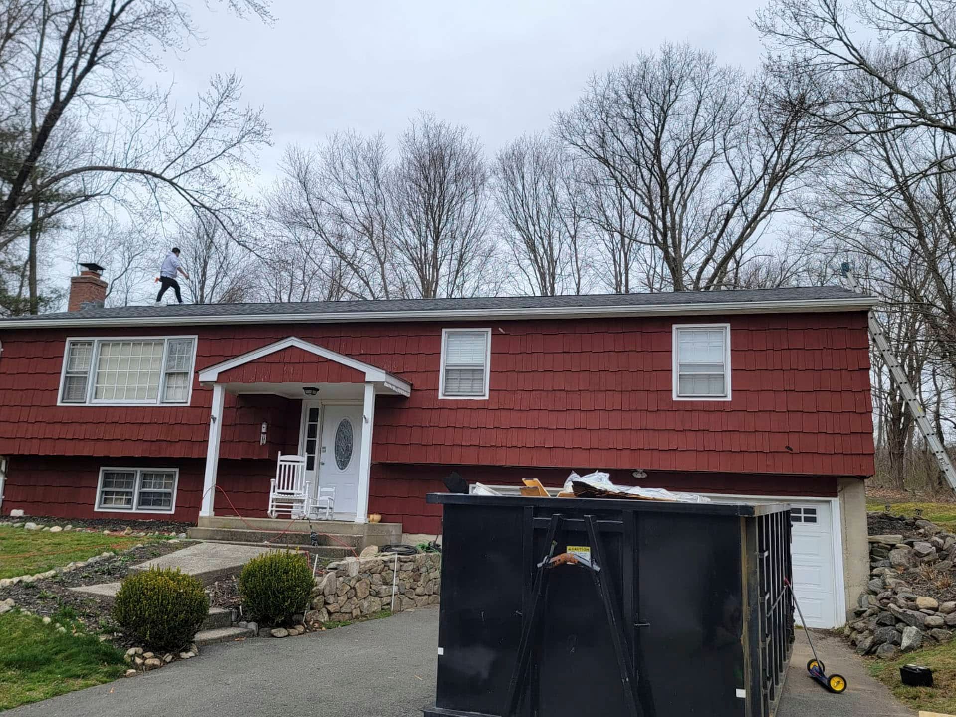 Red house with white trim and porch, viewed from a driveway with two black dumpsters in front.