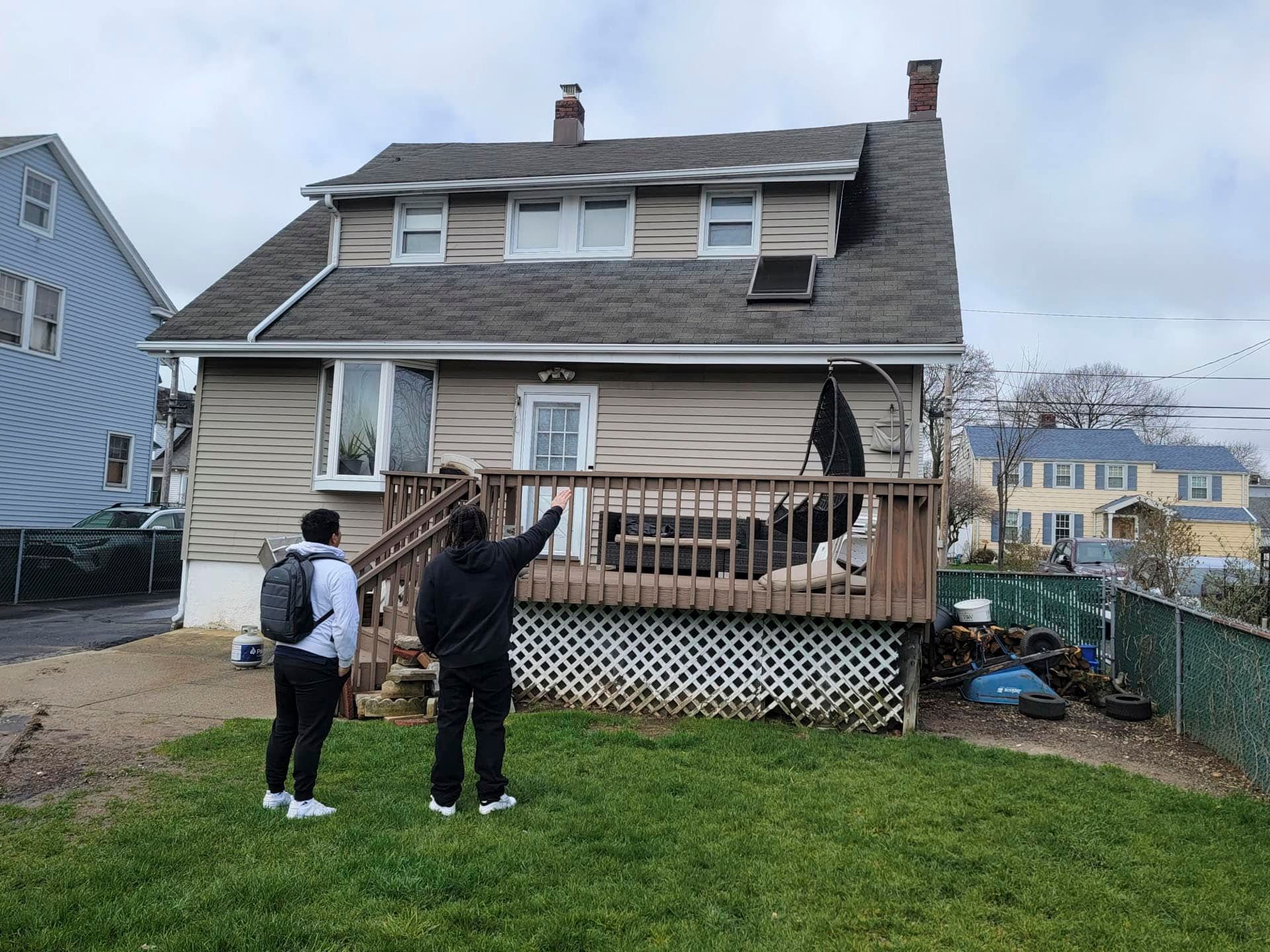 Two people in a yard point toward a house with a covered porch and deck siding.