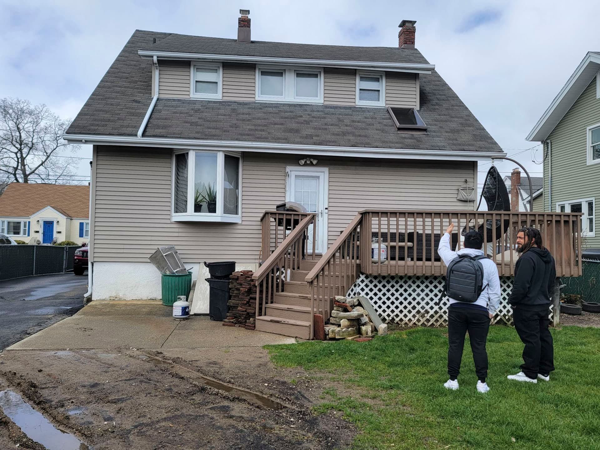 Two people standing in a yard beside a beige house with a raised wooden deck and stairs