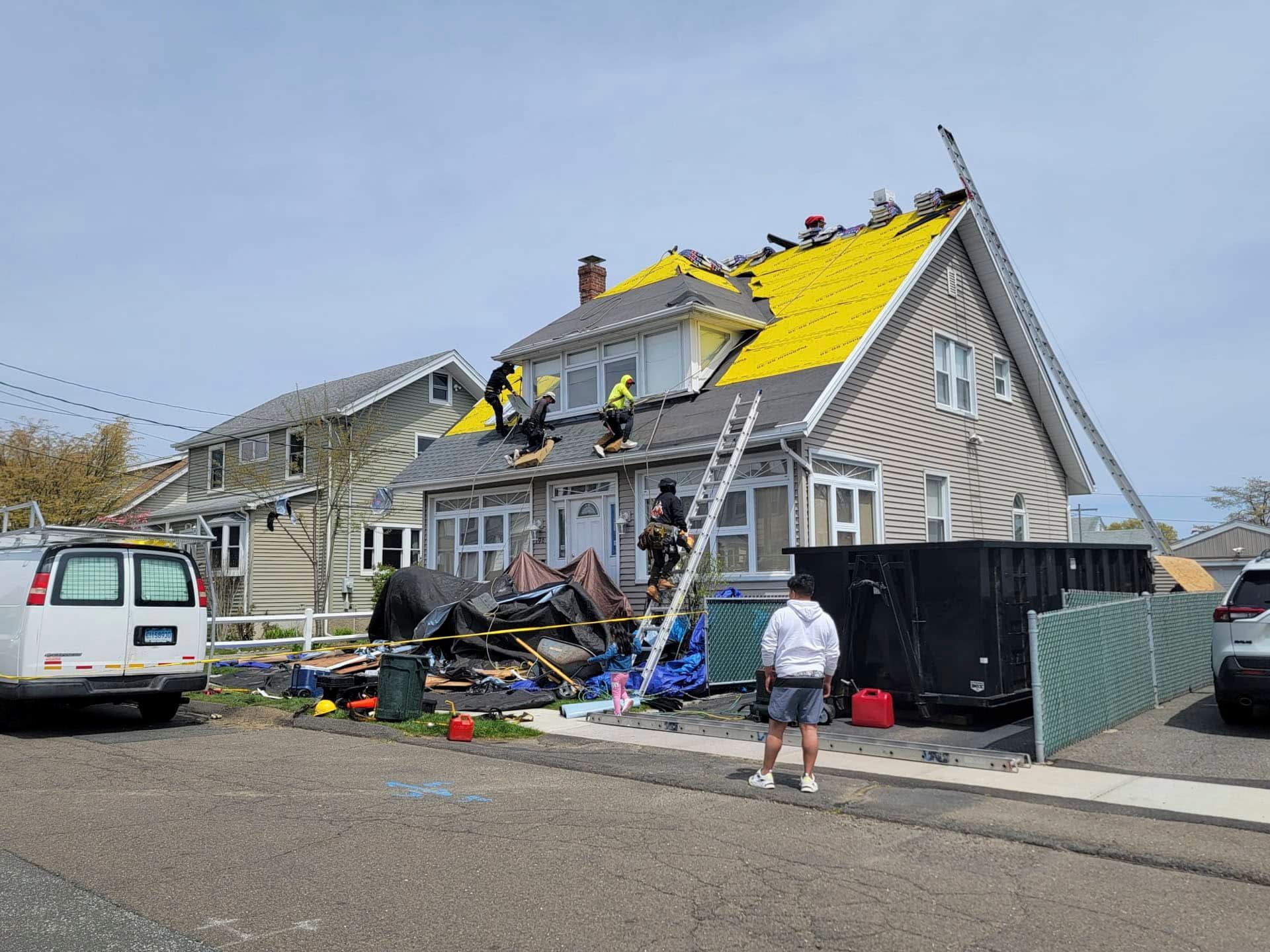 House roof under repair after storm damage, with workers, ladders, and yellow tarps on a residential street