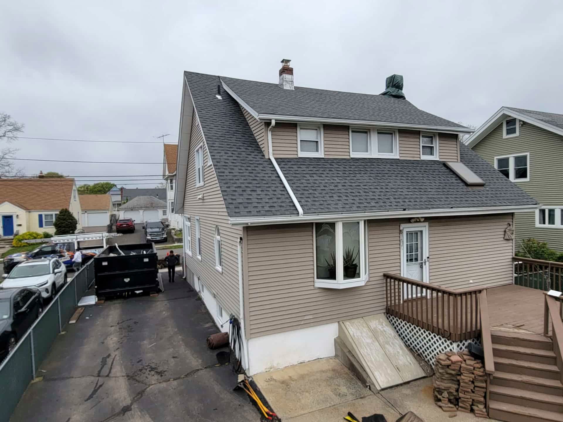 Side view of a beige house with a dark roof, driveway, deck stairs, and parked vehicles on a cloudy day