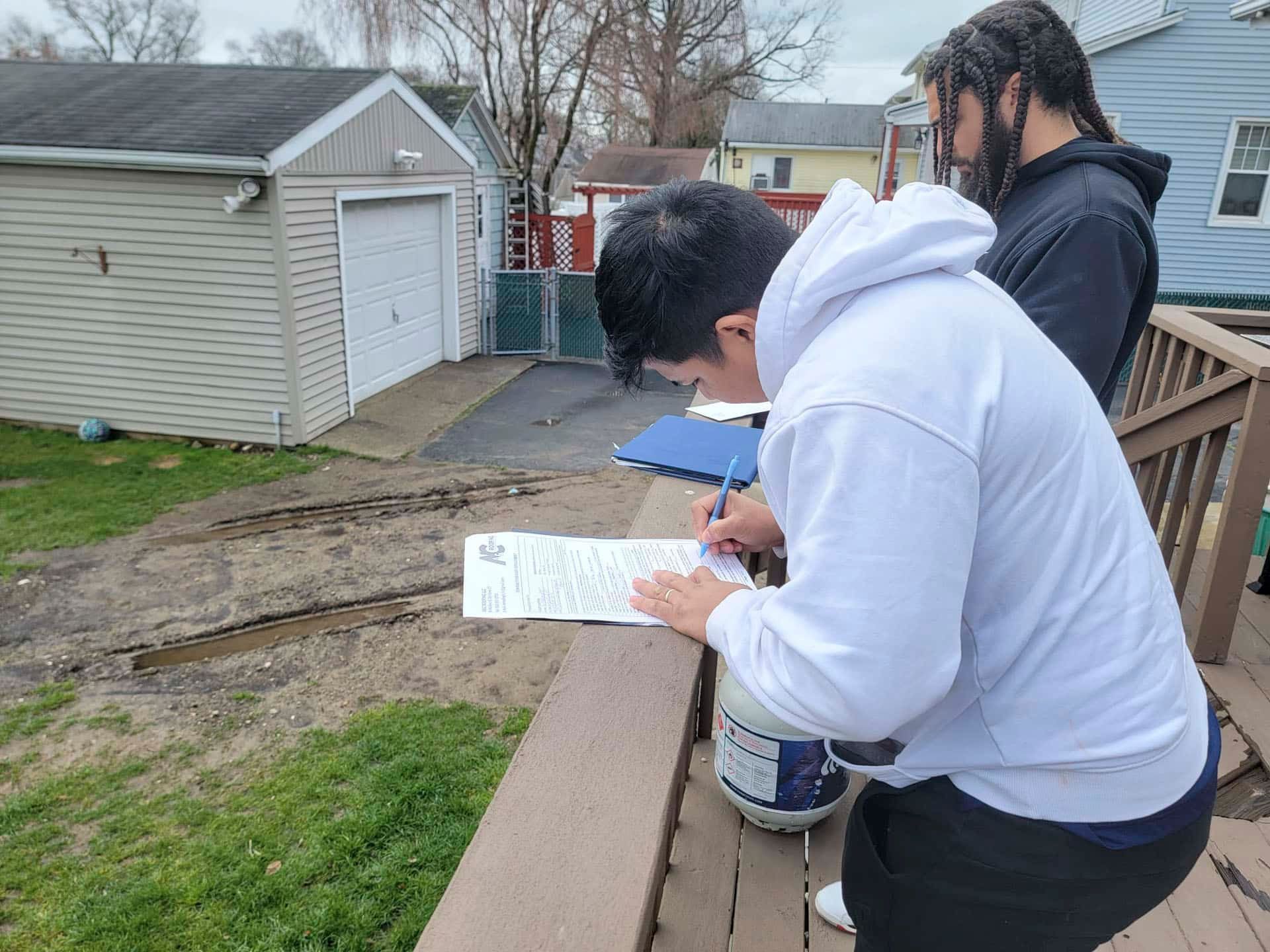 Two people on a deck reviewing papers, with a small shed and muddy yard in the background.