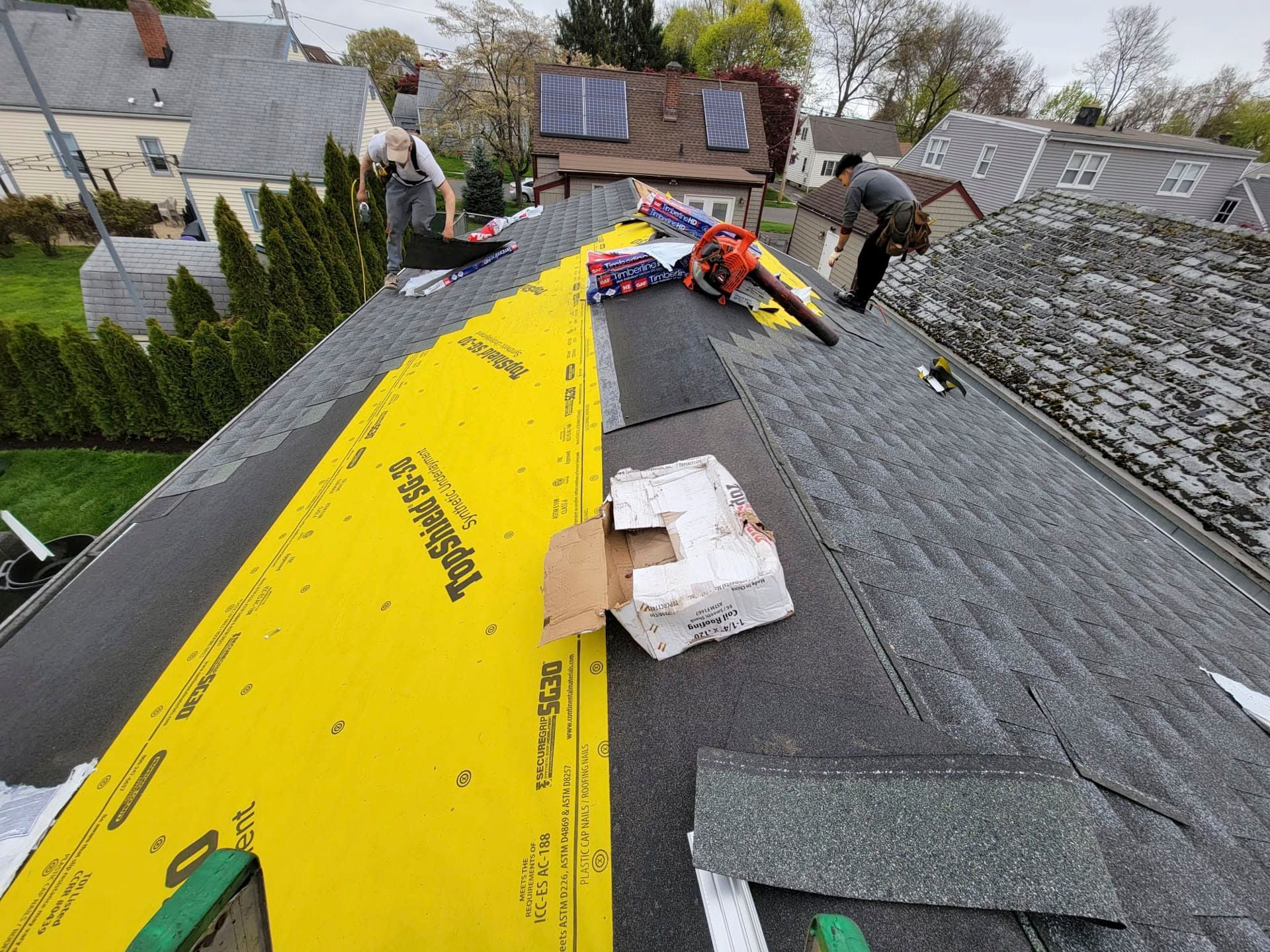 Roofers repairing a shingled roof, with yellow underlayment exposed on a steep rooftop.