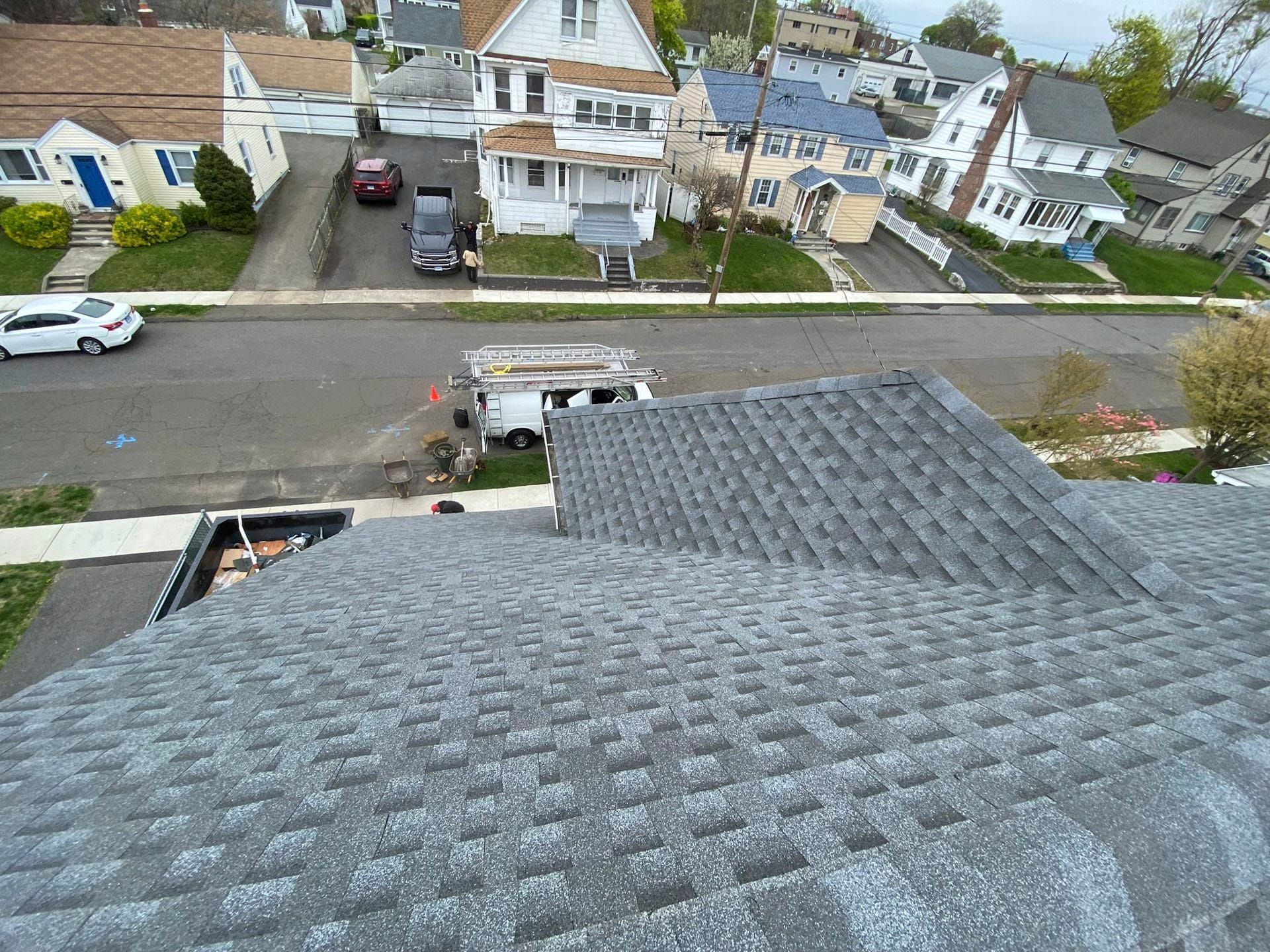 View from a roof over a suburban street with parked cars, houses, and a cloudy sky.