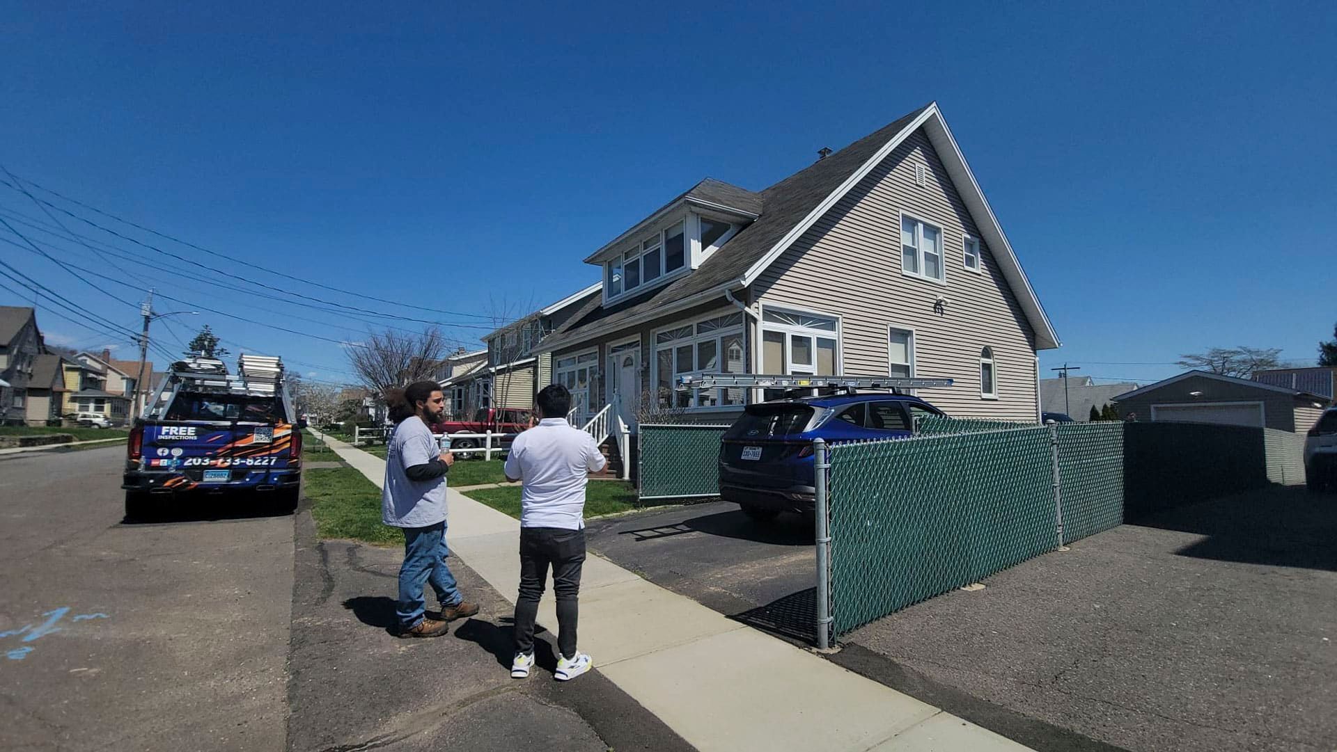 Two people walk past a beige house on a sunny street, with parked cars and a green fence nearby.