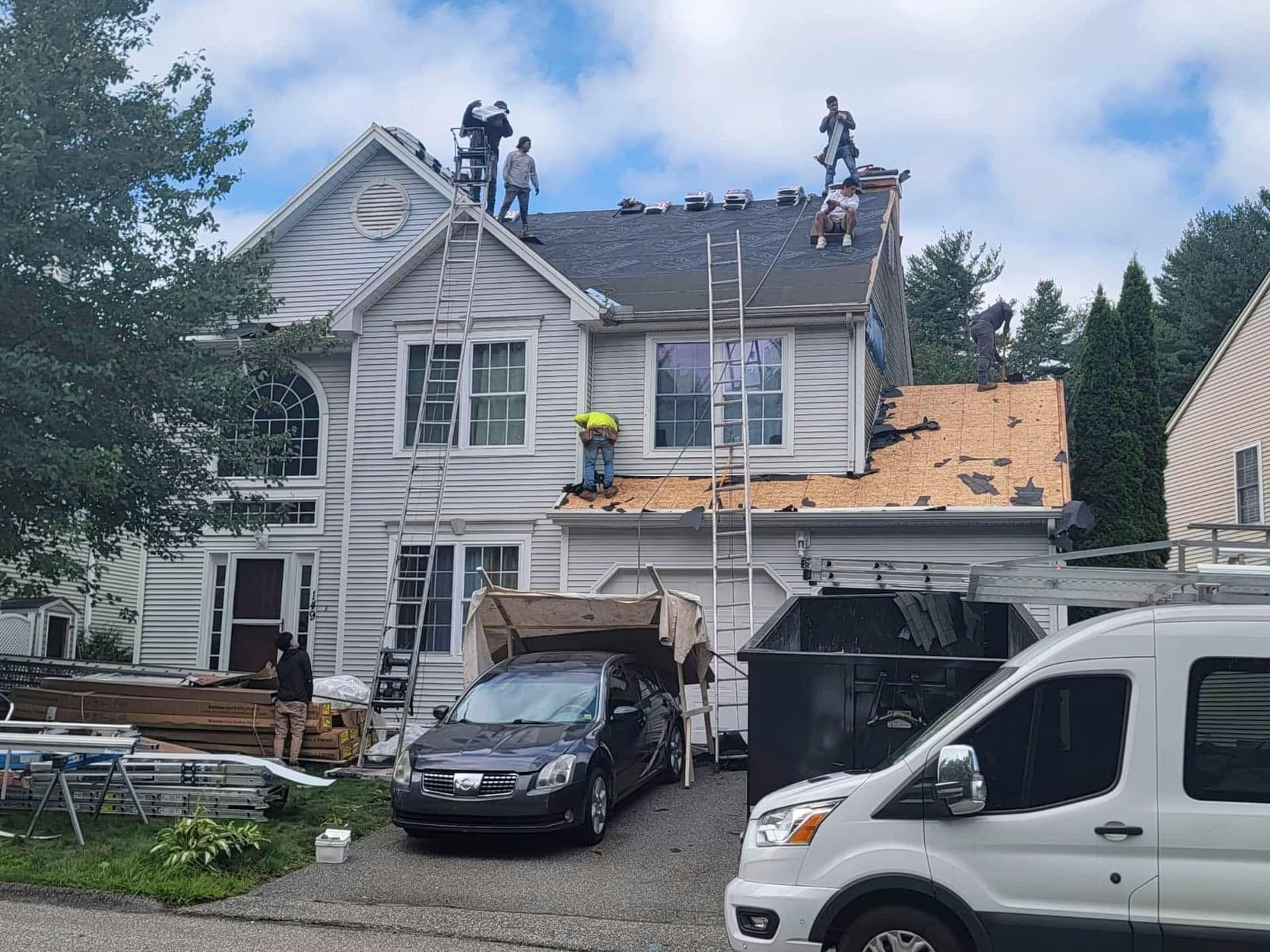 Workers on a roof repairing storm damage on a white house with cars parked in front
