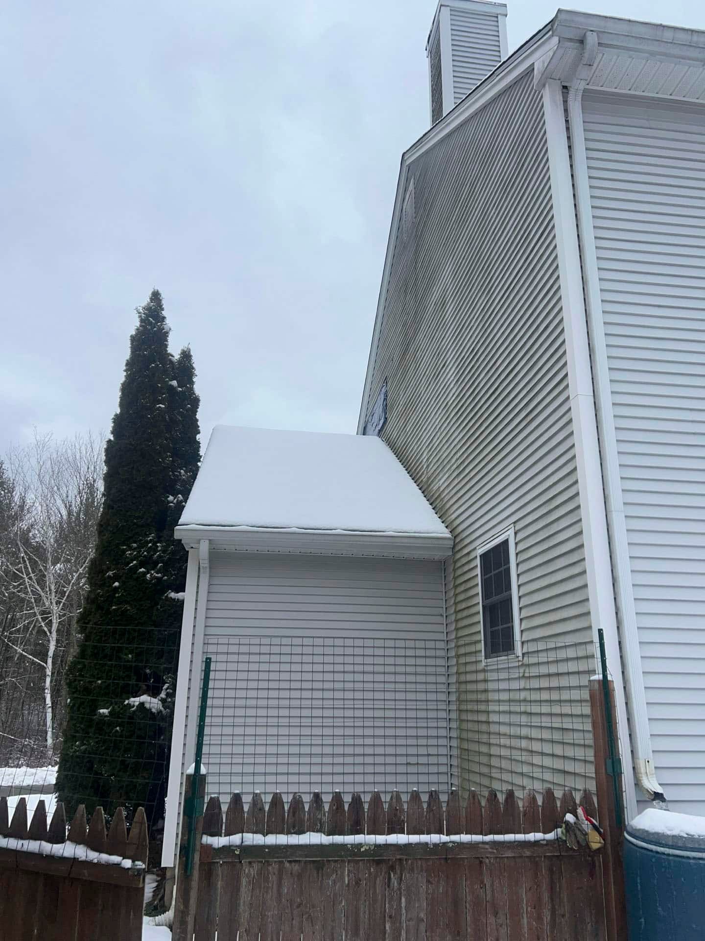 Snow-covered side yard with a white house, tall evergreen tree, and wooden fence on a cloudy day