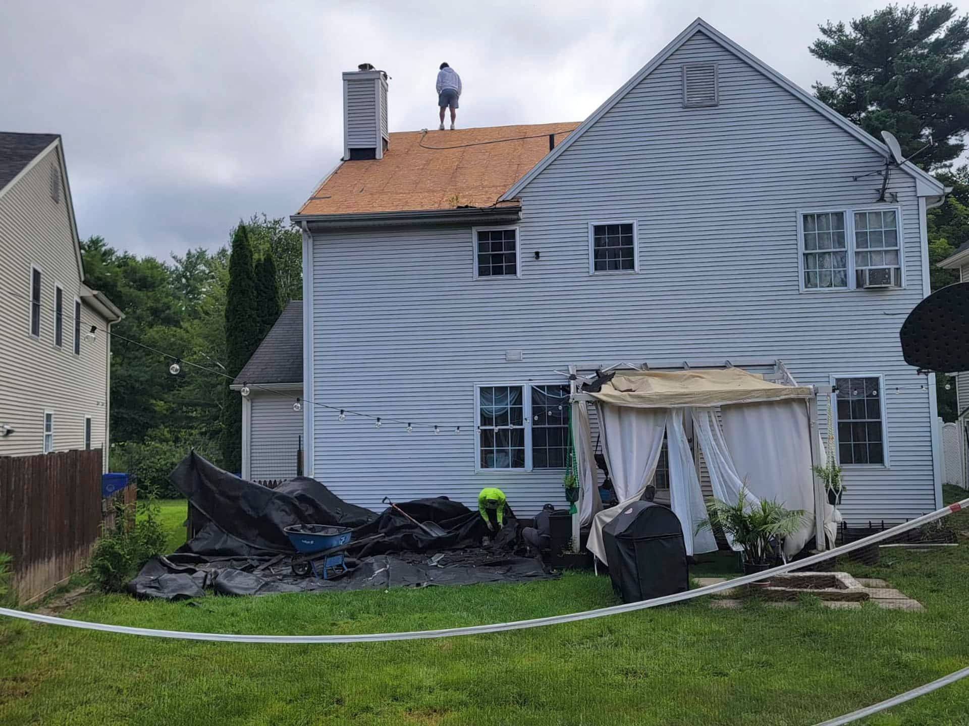 Damaged house with tarp-covered debris in yard and a worker on the roof after storm damage
