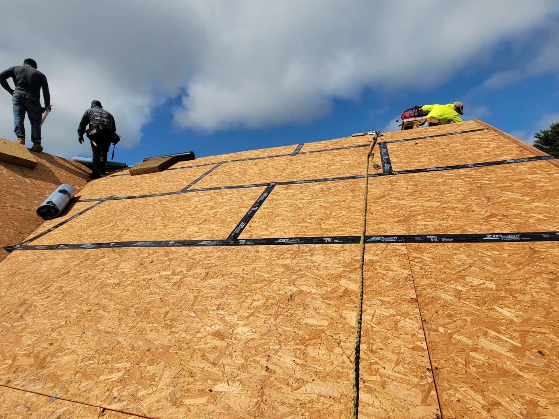 Workers installing plywood sheathing on a steep roof under a cloudy sky