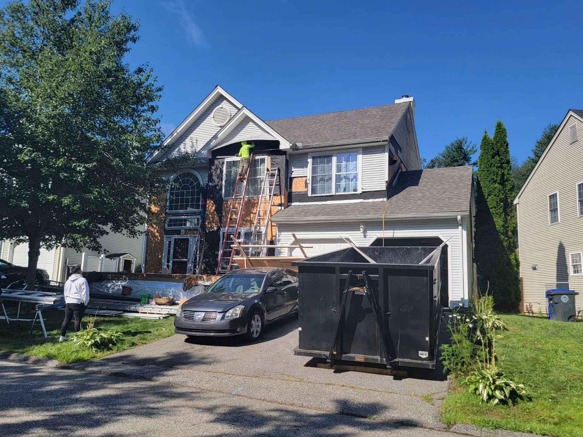 House with workers on front porch and a car in the driveway, with a large dumpster in front