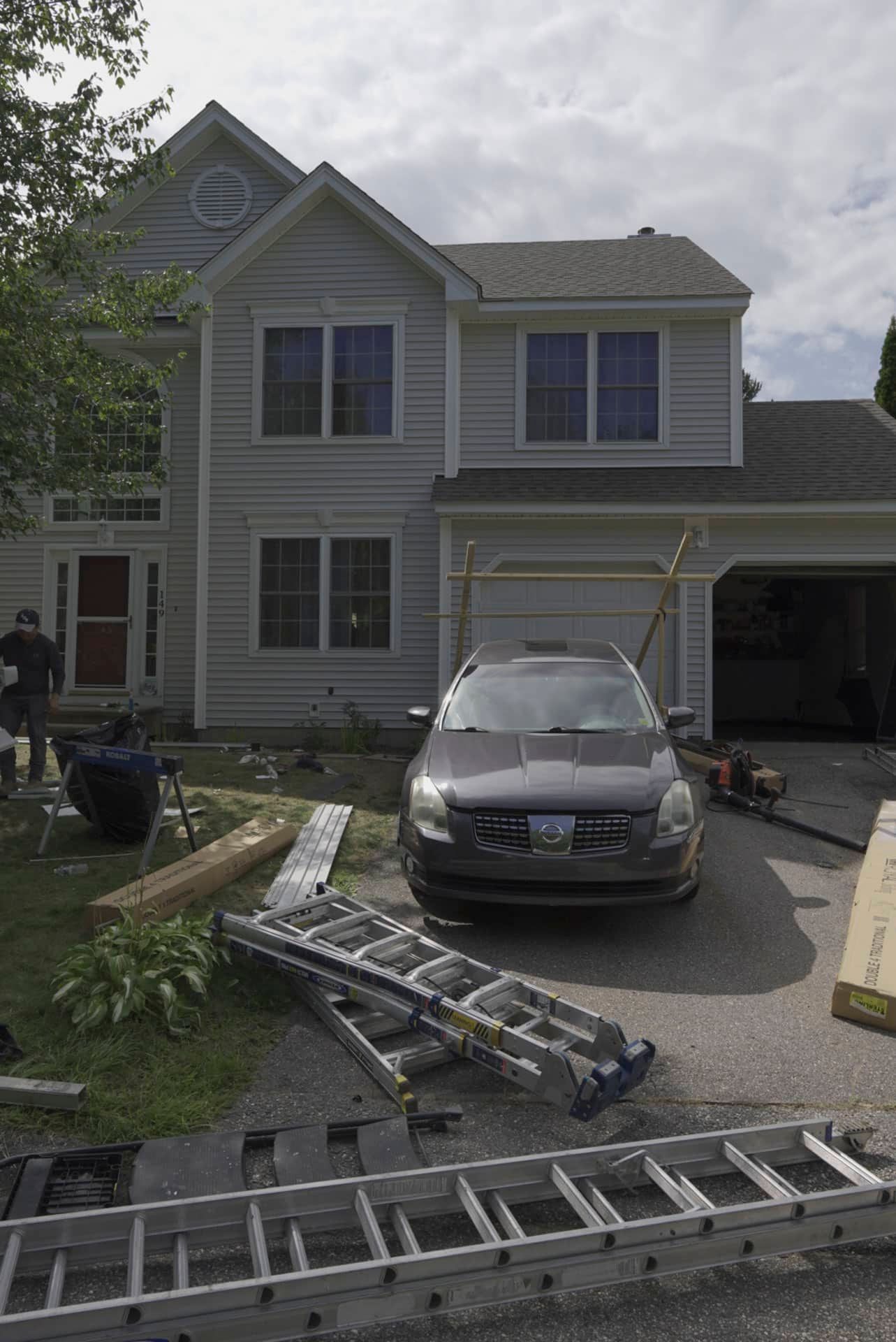 Two-story house with car in driveway, ladders and tools scattered on front lawn and walkway, cloudy day