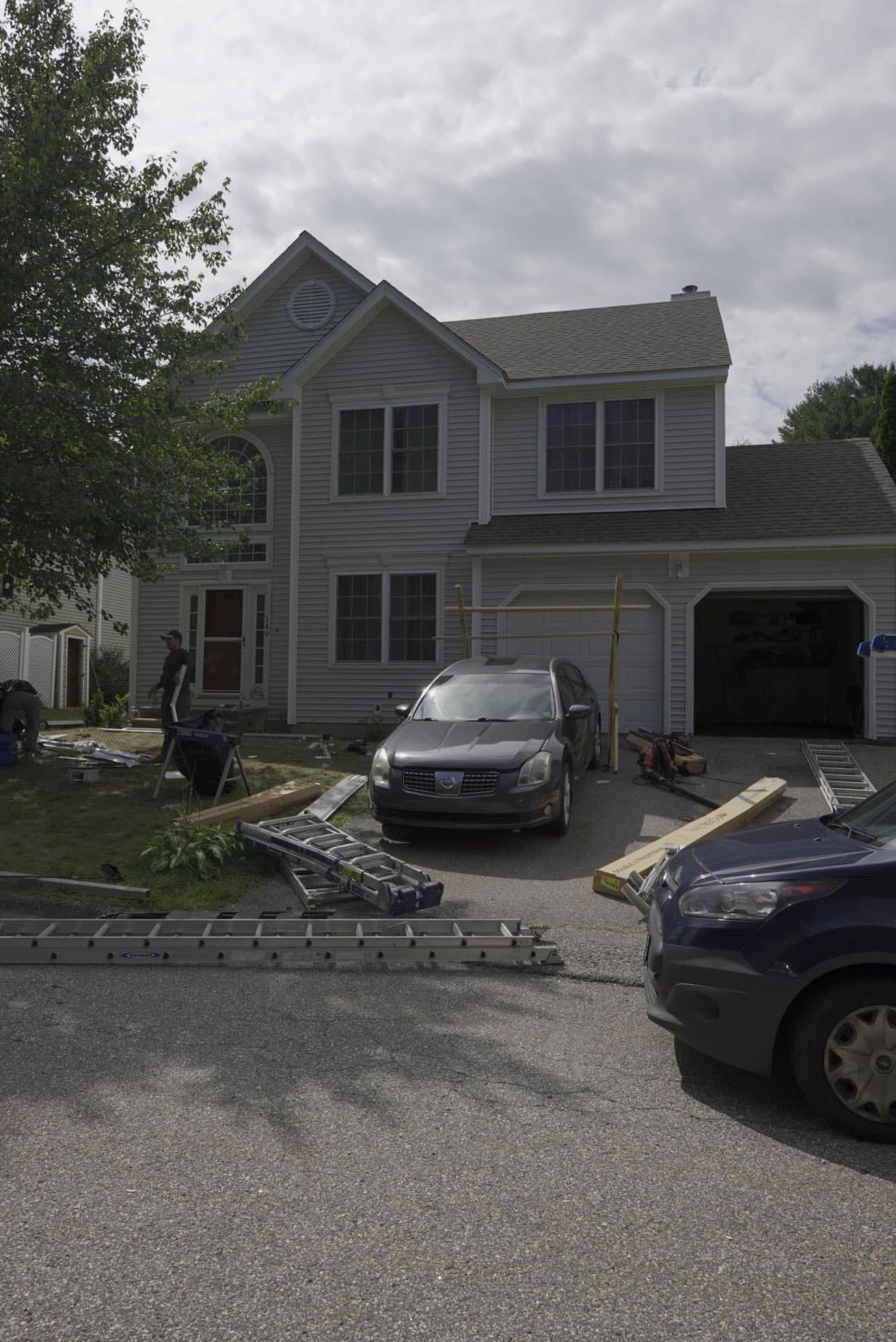 Damaged suburban house with debris in driveway and a car parked outside under cloudy skies