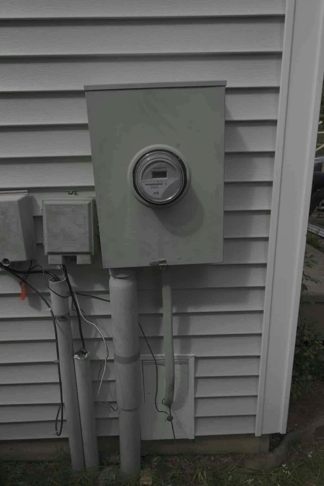 Exterior electrical meter and service boxes mounted on a gray house wall.