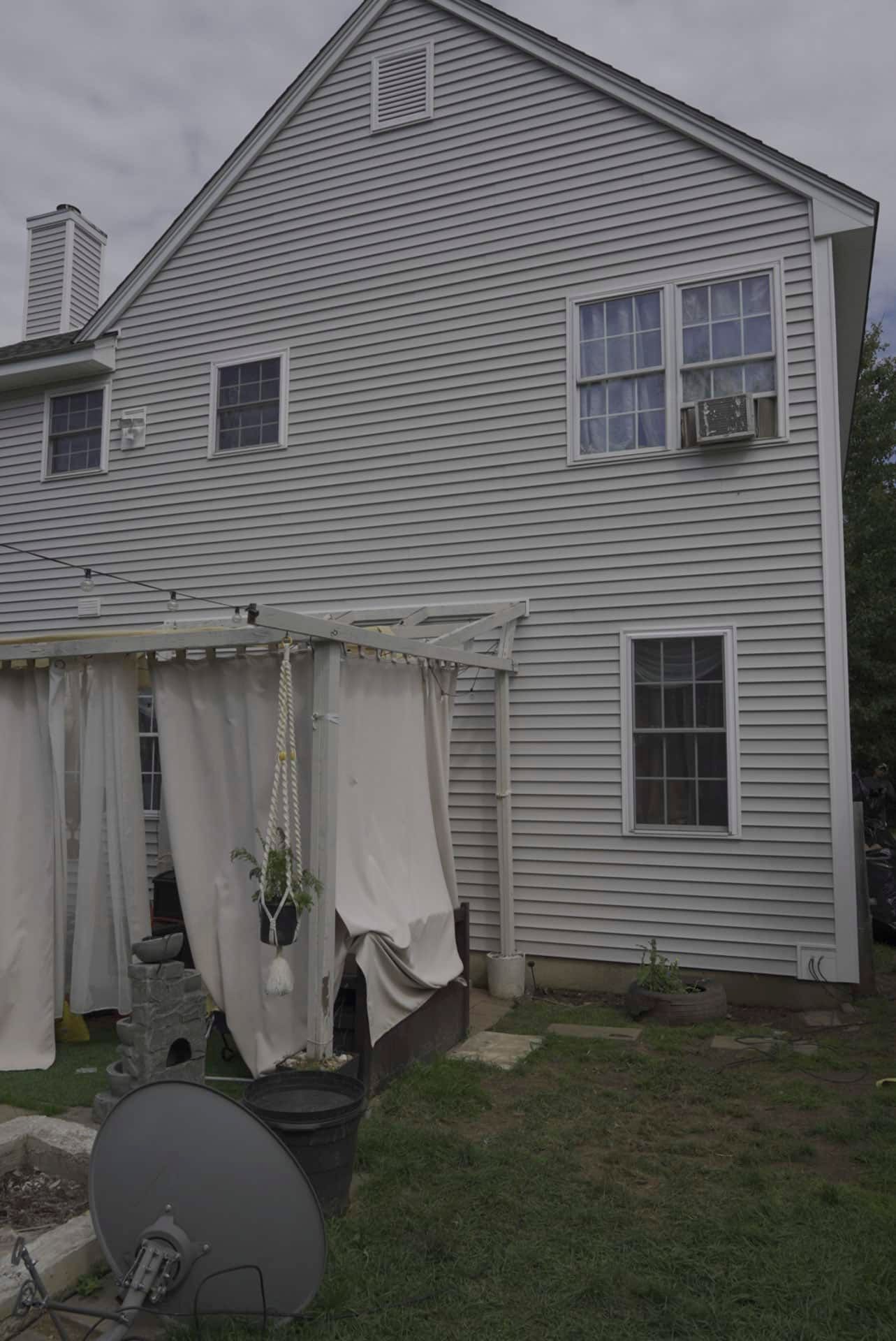 Back of a gray house with a covered patio, curtains, satellite dish, and lawn