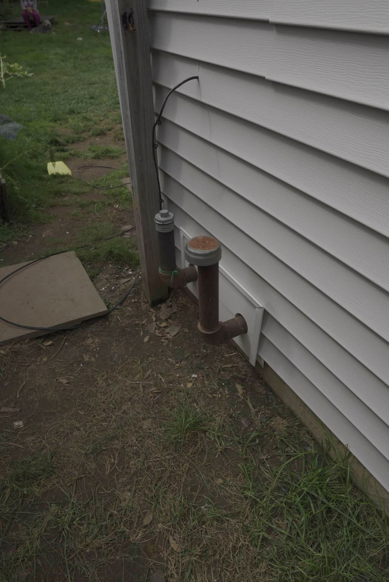 Utility pipes and vent along a house’s gray siding beside a grassy yard.