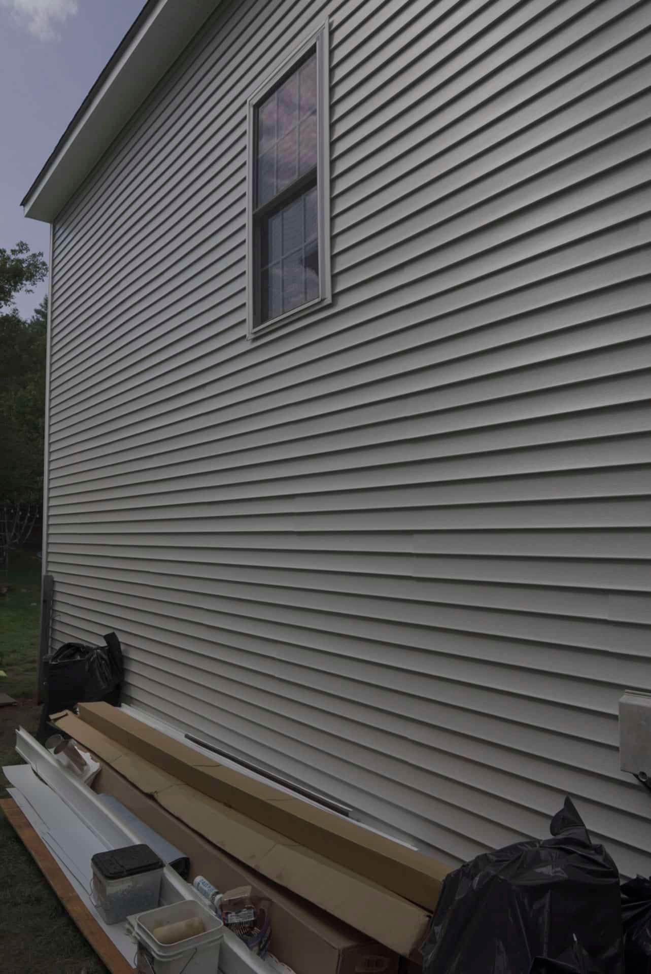 Exterior house wall with beige vinyl siding, a window, and construction materials stacked along the foundation