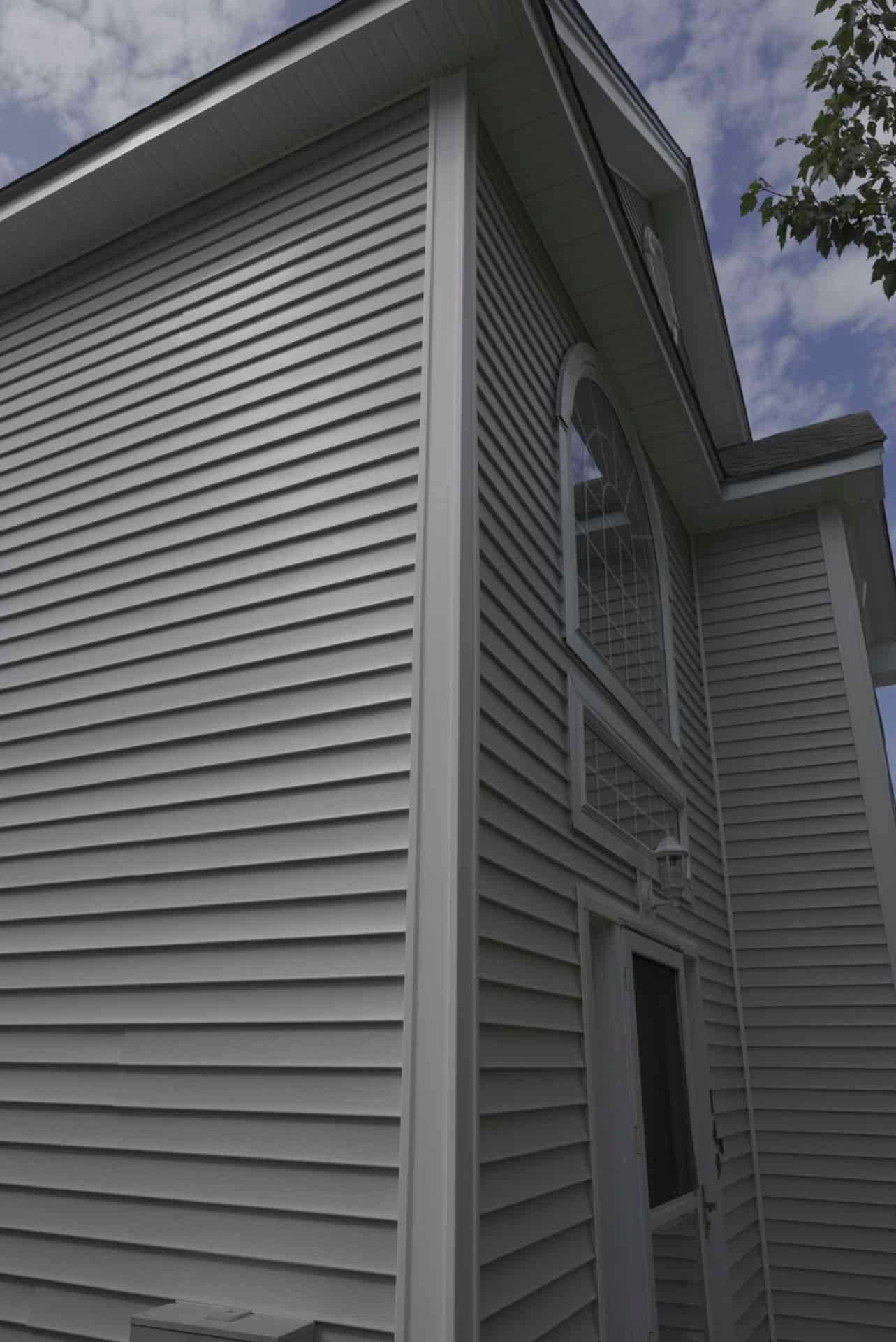 Exterior corner of a gray vinyl-sided house with white trim and a visible window under the roofline