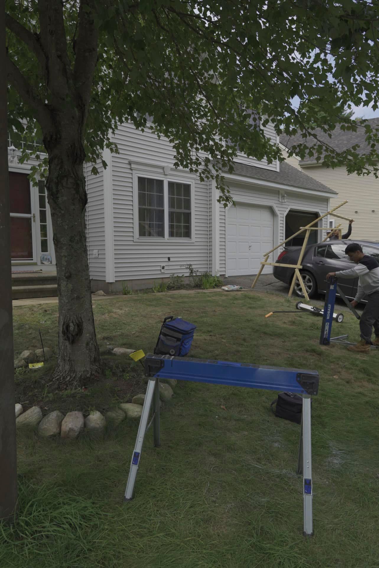 Blue folding chair in a grassy yard with a tree, house, and people working near a truck in the background