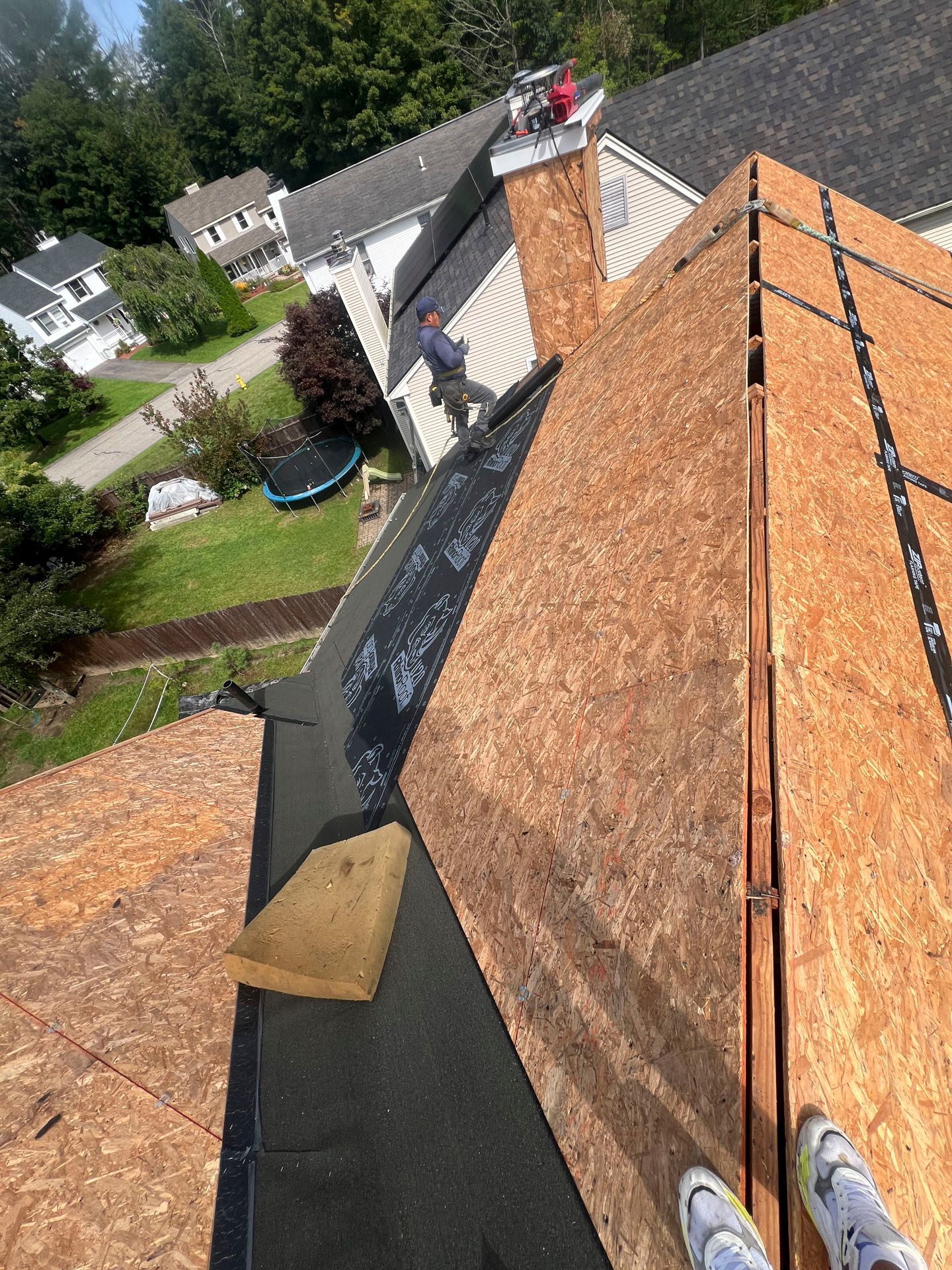 Person standing on a steep shingled roof with a chimney and suburban houses below.