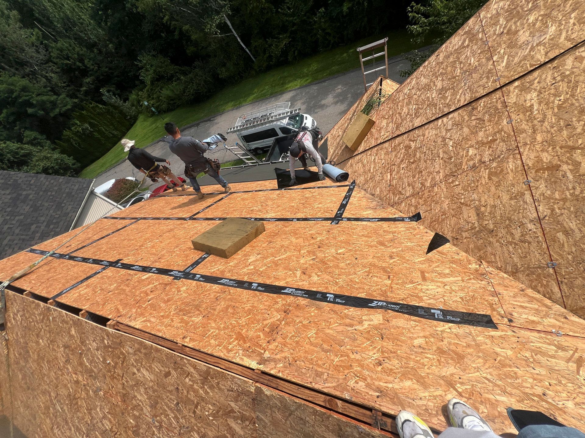 Roofers installing plywood sheathing on a steep roof, with tools and materials on the roof edge.