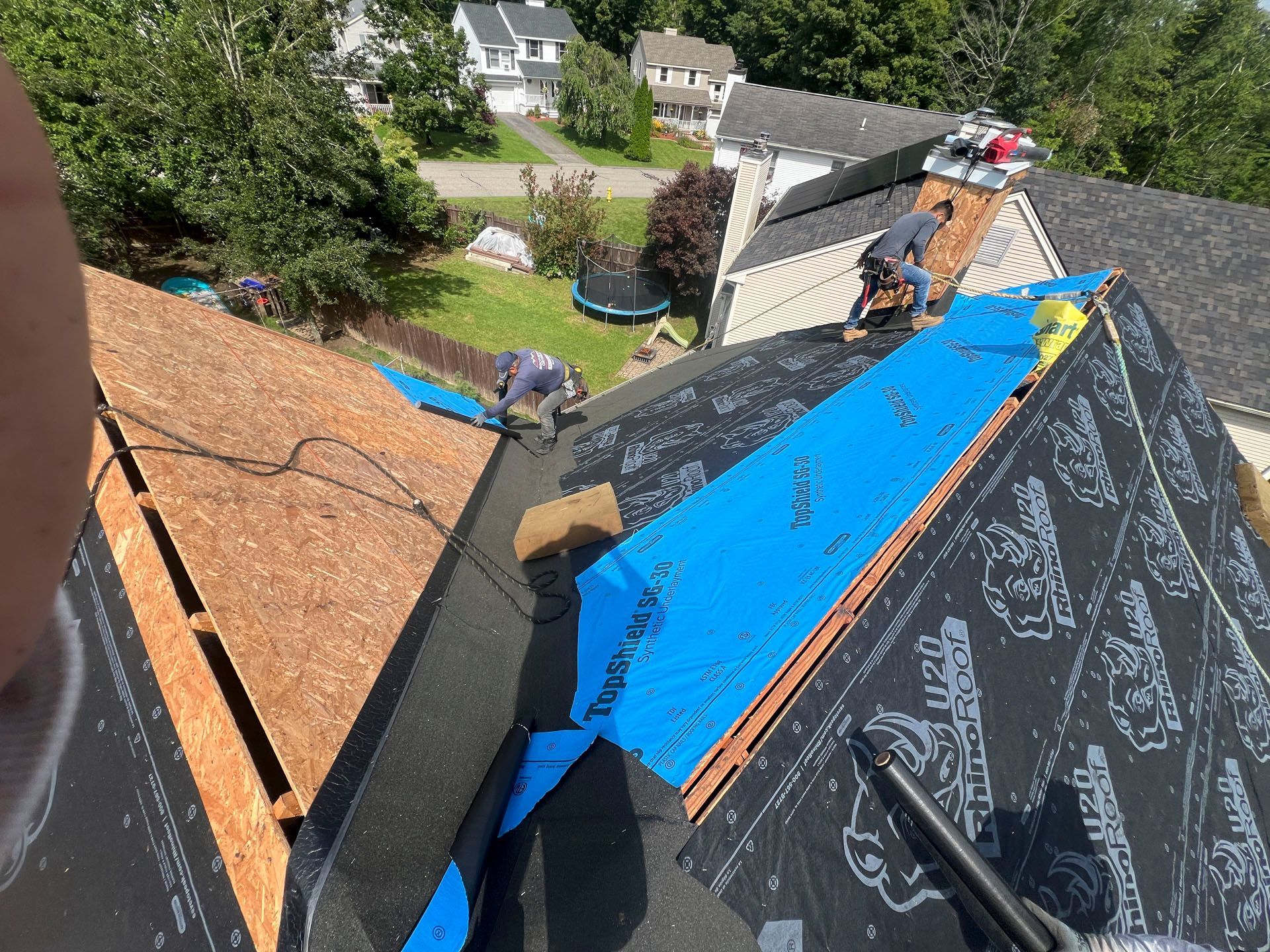 Roofers installing blue underlayment on a steep shingle roof near a residential backyard