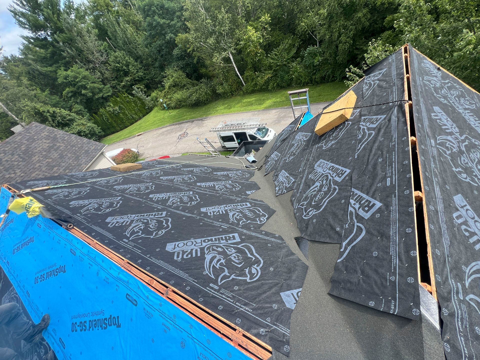 Roof under construction with black underlayment, blue tarp, and trees in the background
