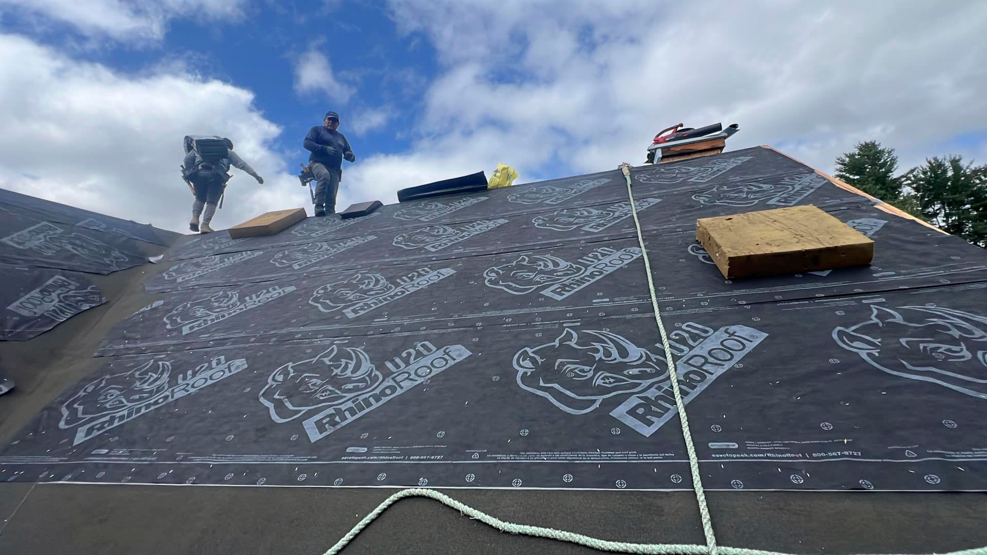 Roofers installing black underlayment on a sloped roof, with tools and materials spread out.