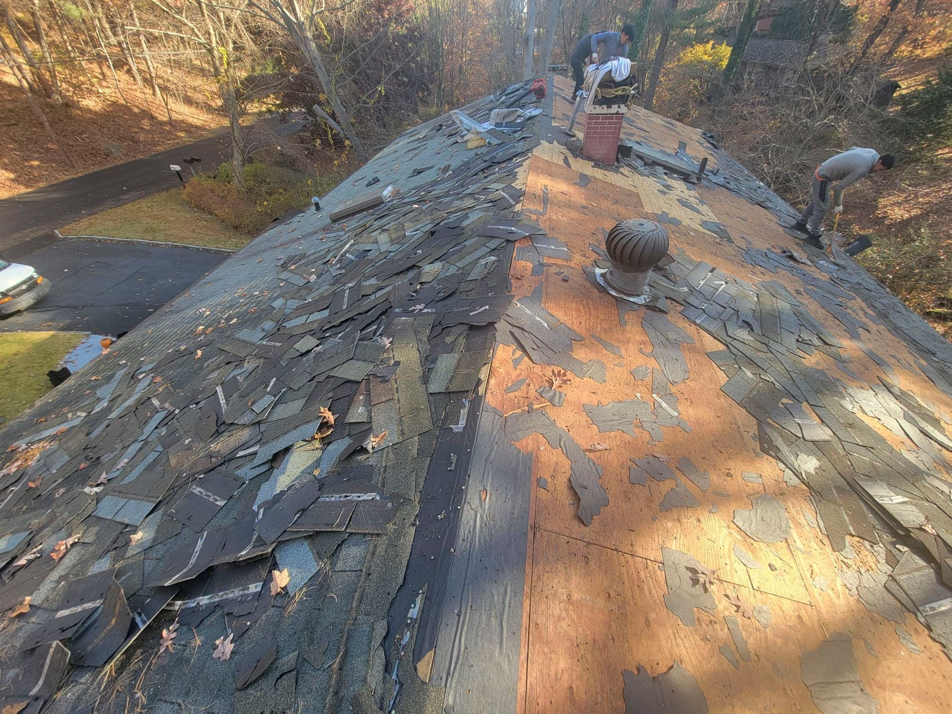 Damaged roof with missing shingles and exposed plywood on a house in a wooded area