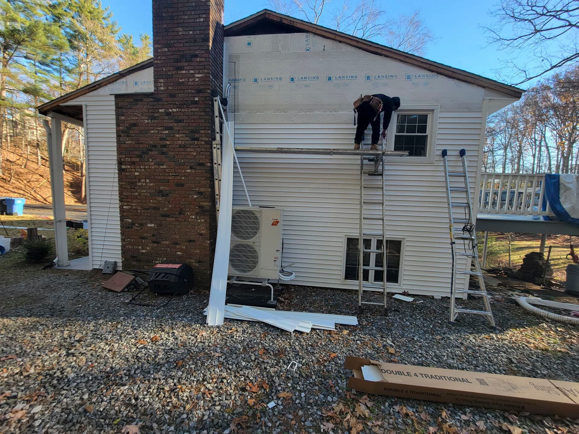 Worker on a ladder repairing white siding on a house exterior with brick chimney.