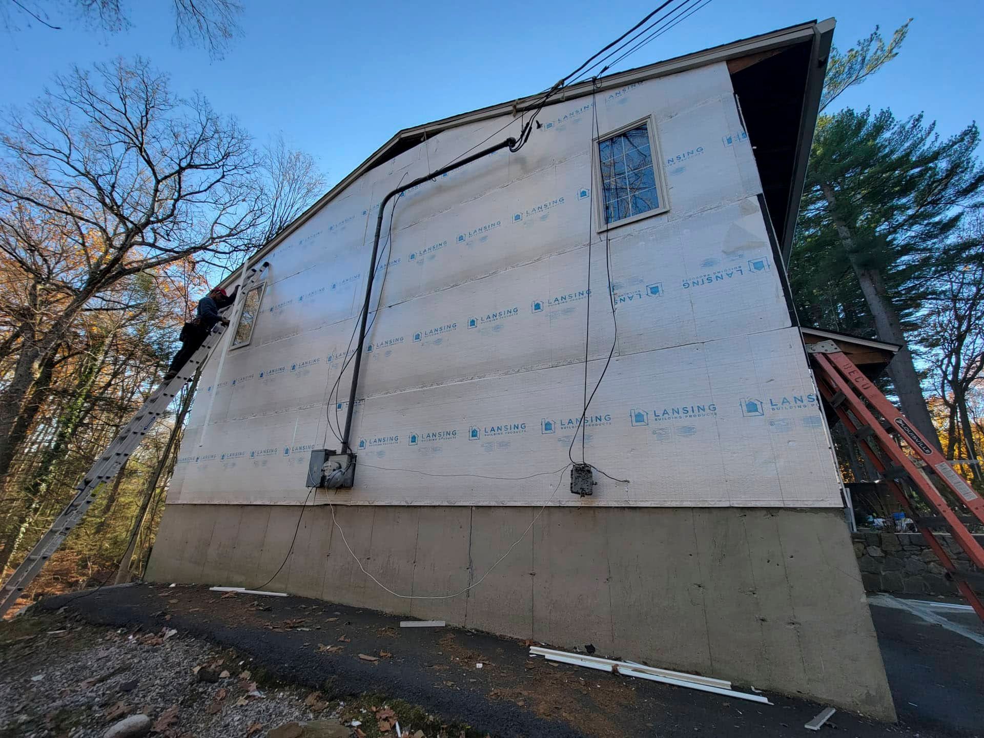 Exterior of a house wrapped in white weatherproofing, viewed from below with trees and a ladder nearby