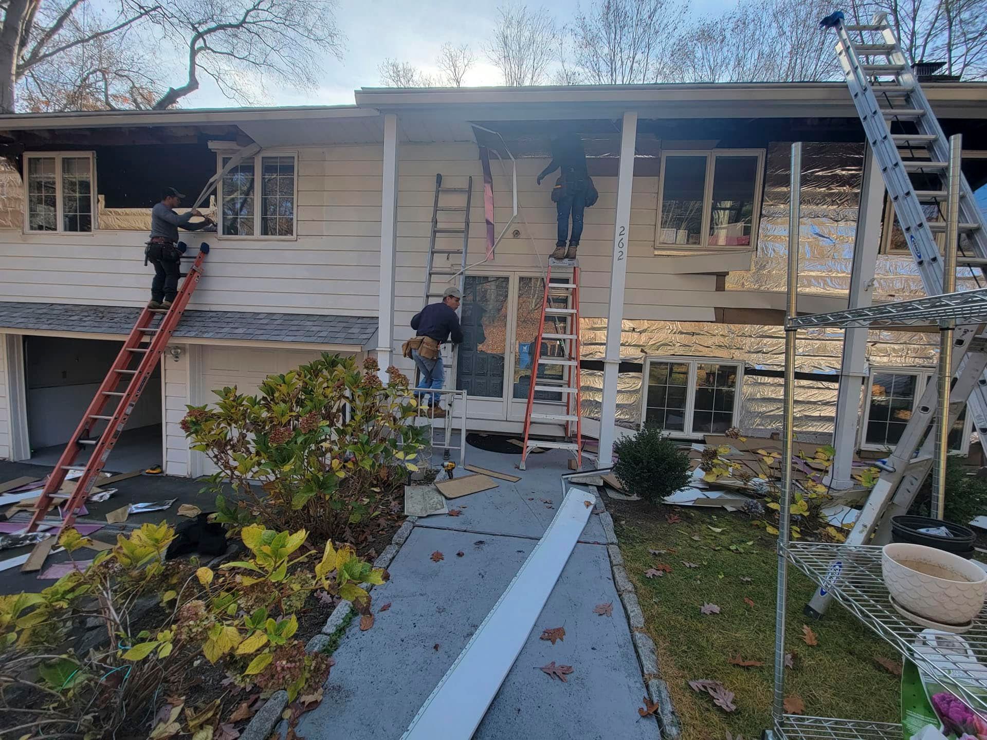 Workers on ladders repairing the roof and siding of a house with a stone front.