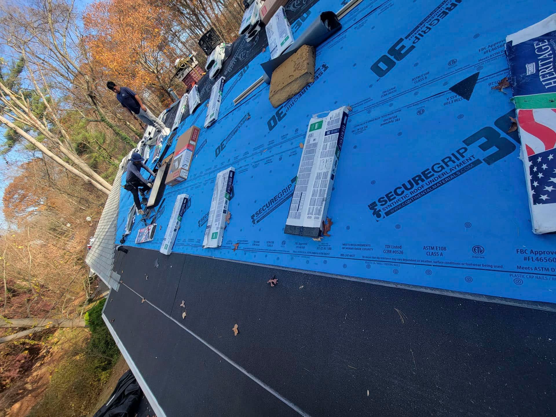 Roof under construction with blue underlayment, scattered materials, and trees in the background.
