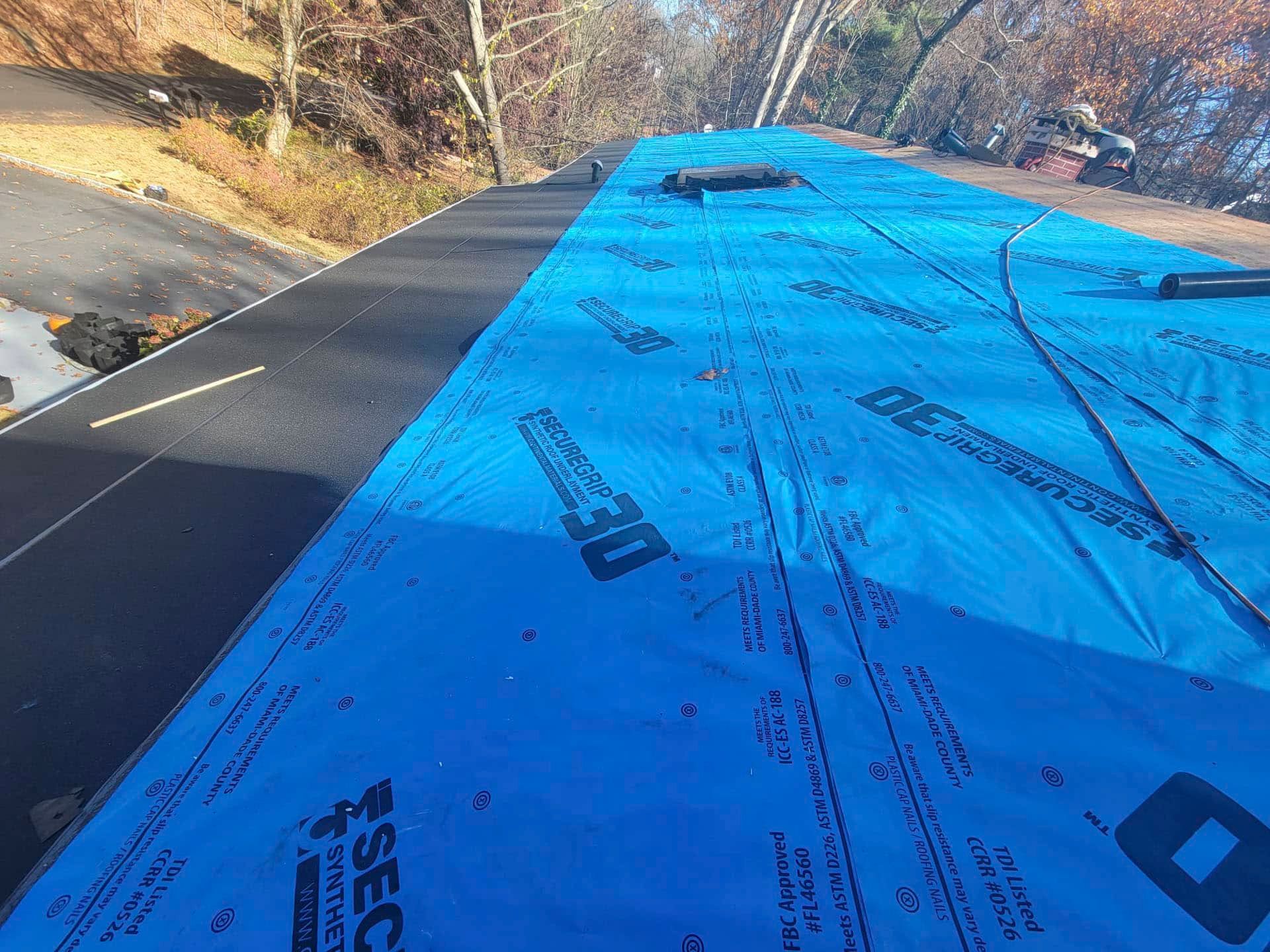 Blue roofing underlayment covering a sloped roof beside trees and a driveway
