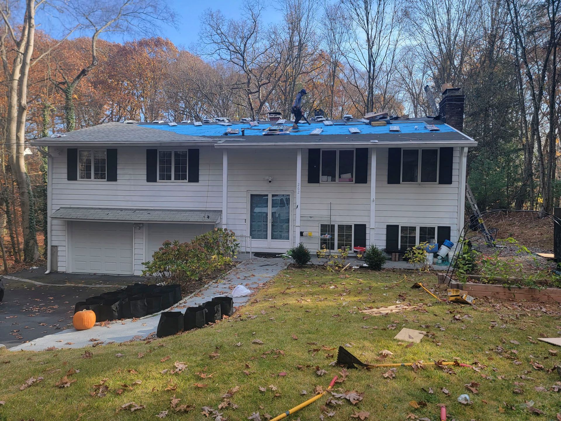 Two-story white house with blue tarp on roof and damaged siding, surrounded by trees and a grassy yard
