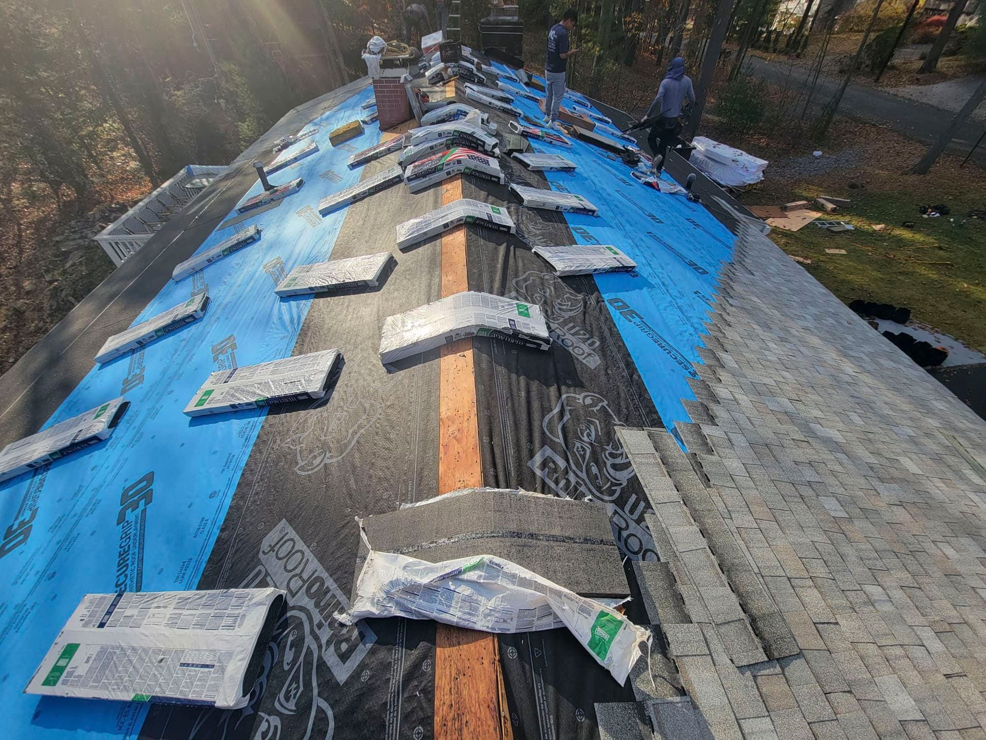 Roof under repair with blue tarp, gray shingles, and ridge vent in a wooded area