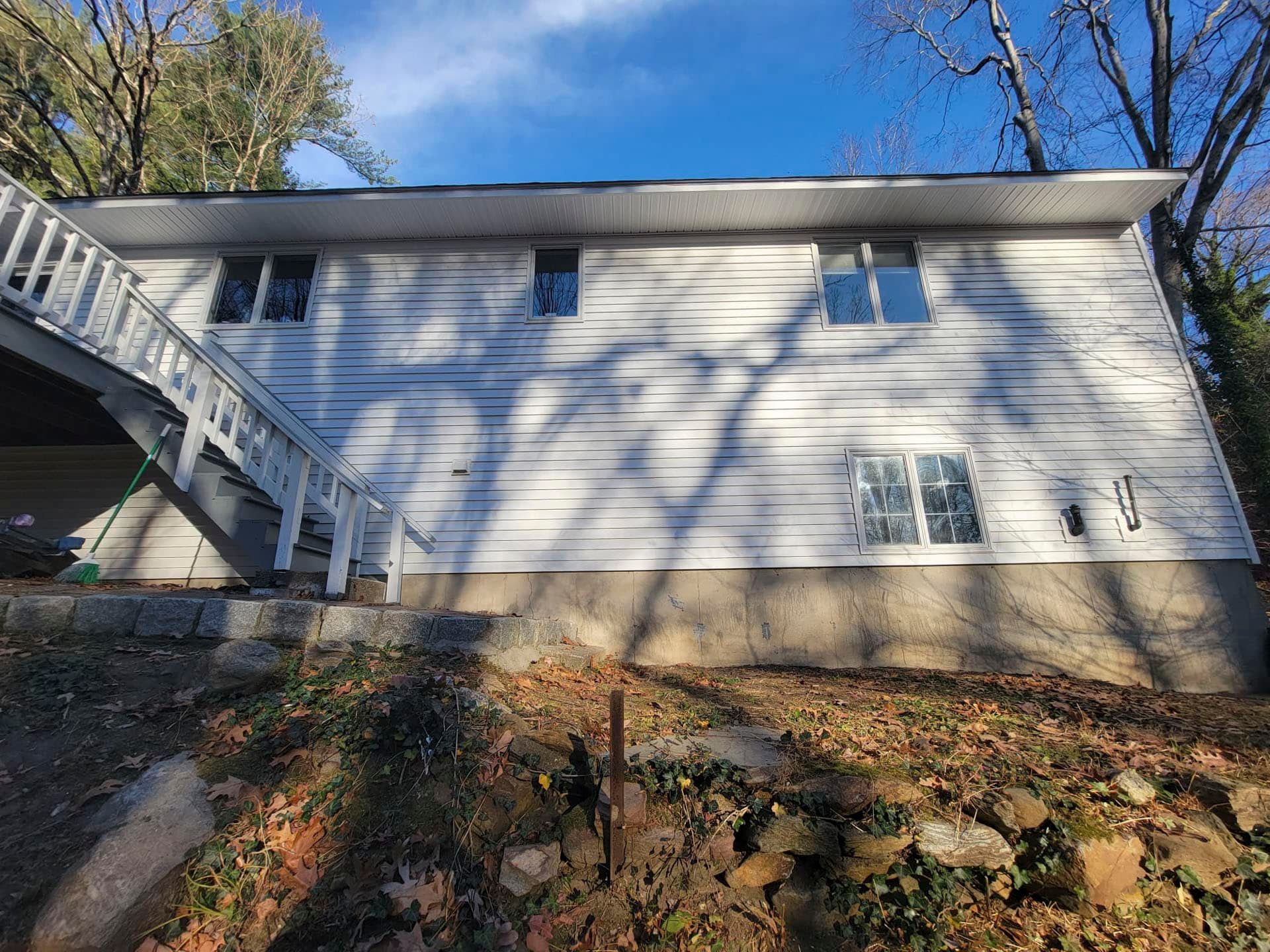 Side view of a white house with an exterior staircase, beige foundation, and trees casting shadows.