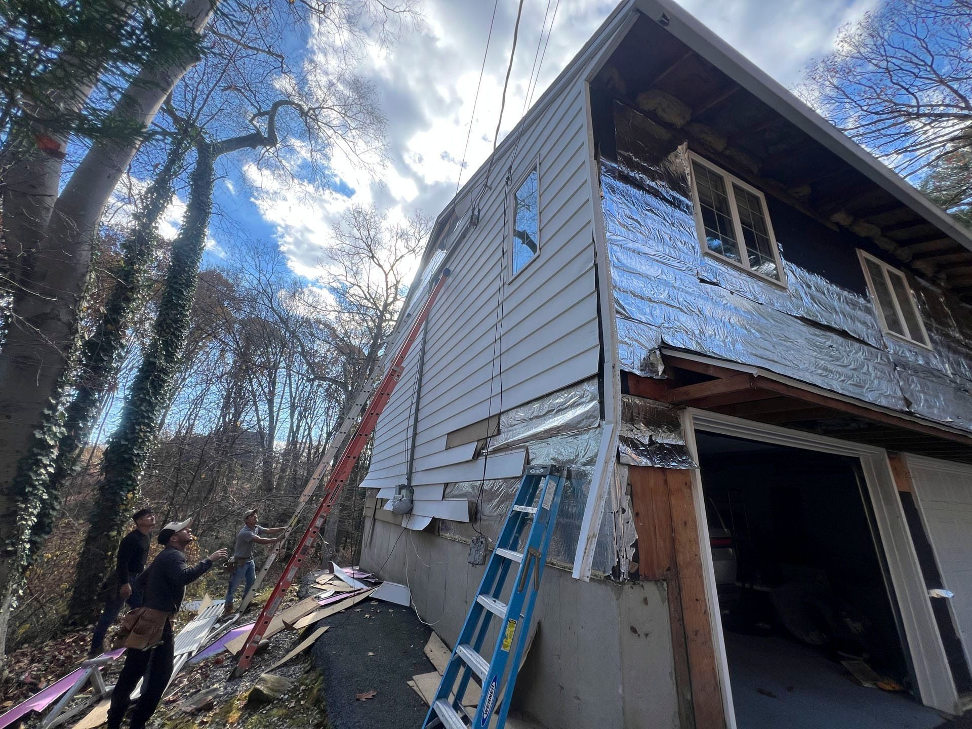 Two-story house under repair with ladders leaning against the siding and people working outside