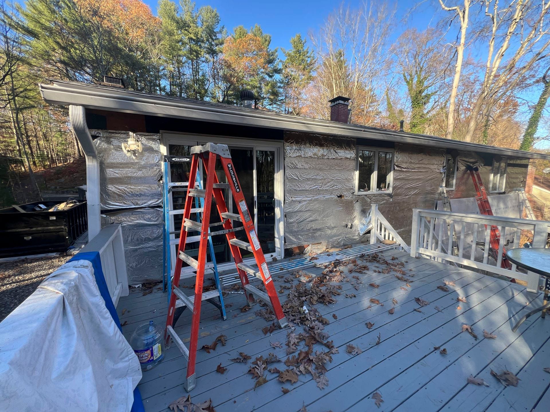 Ladder leaning against a house with a deck, autumn trees, and construction materials on the roof.
