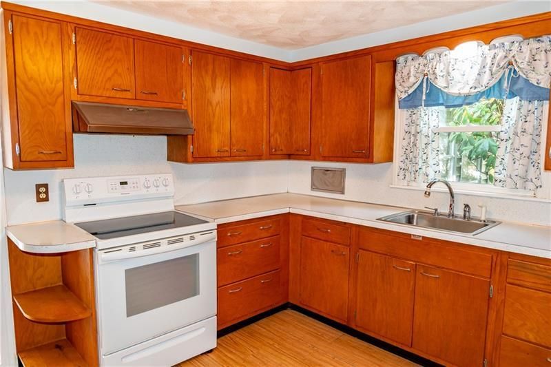 Kitchen with wood cabinets, white stove, and sink. Window with floral curtains.