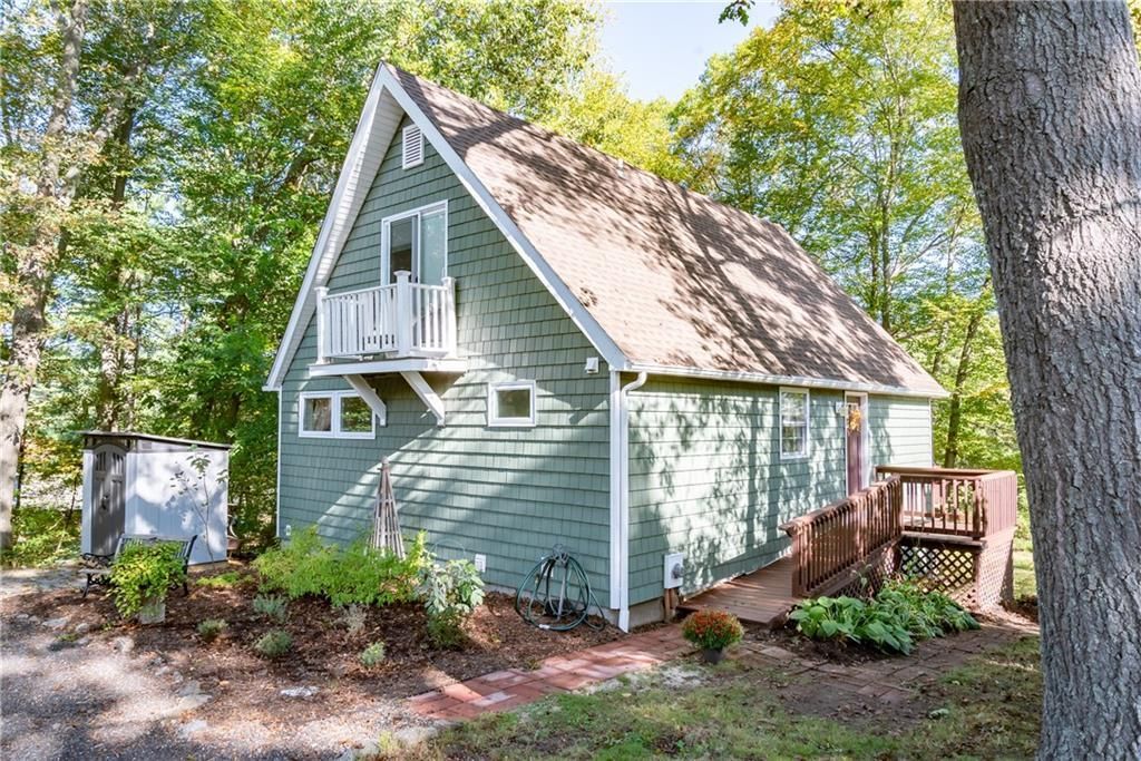 Green cottage with a balcony and a brown roof surrounded by trees.